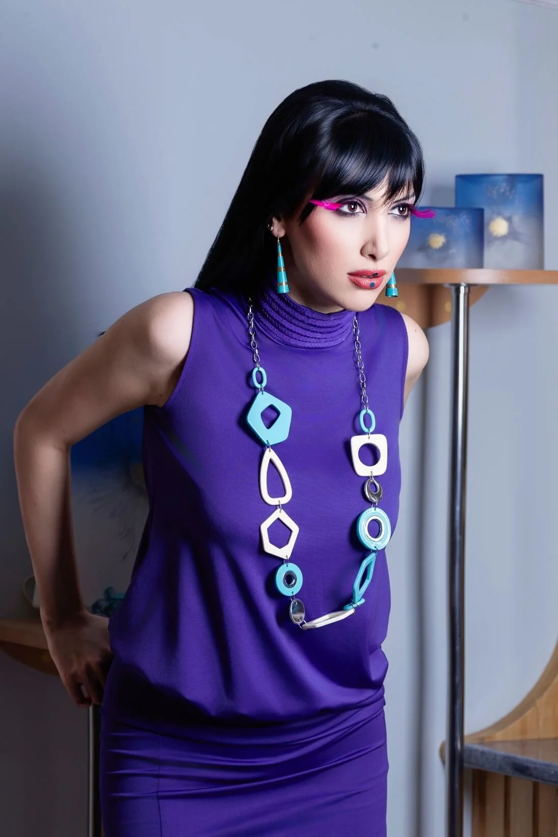 A woman with black hair and bright pink glasses wearing a purple sleeveless dress and a large blue and white geometric statement necklace, standing in a modern room.