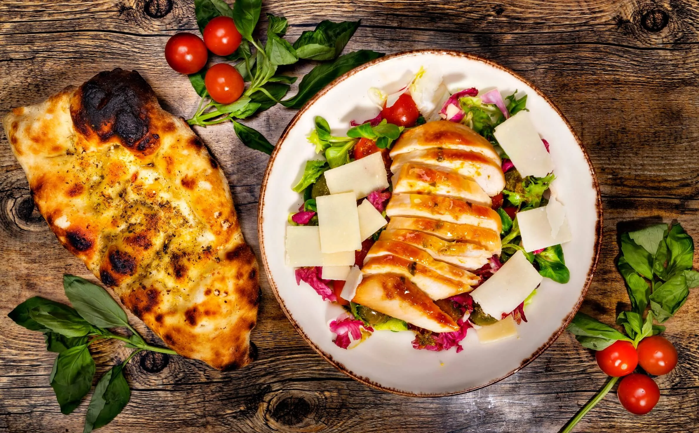 A wooden table holding a slice of cheese pizza on the left and a salad with grilled chicken, cherry tomatoes, leafy greens, cherry tomatoes and parmesan cheese on a white plate at the center, with additional cherry tomatoes and basil leaves scattered