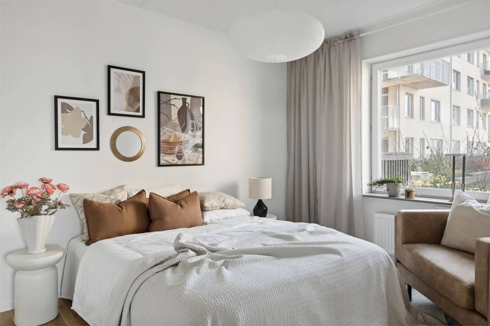 A cozy bedroom with a white bed and brown pillows, a white bedside table with pink flowers, a window with beige curtains, and wall art above the bed, including framed pictures and a round mirror, with a tan armchair near the window.