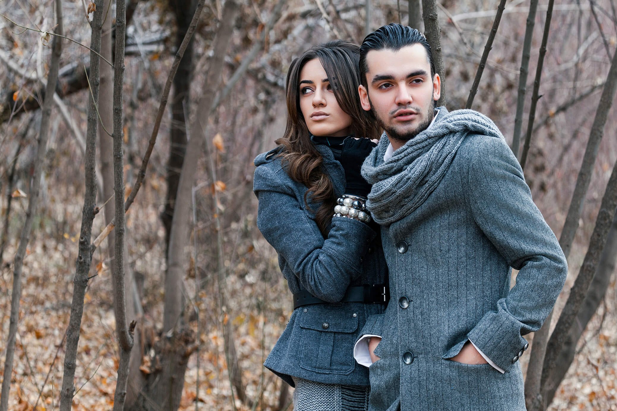 A young man and woman standing close together outdoors in a wooded area with bare trees and fallen leaves. The woman has long brown hair, and the man has dark hair slicked back. They are both wearing gray coats, and the woman has a black scarf and la