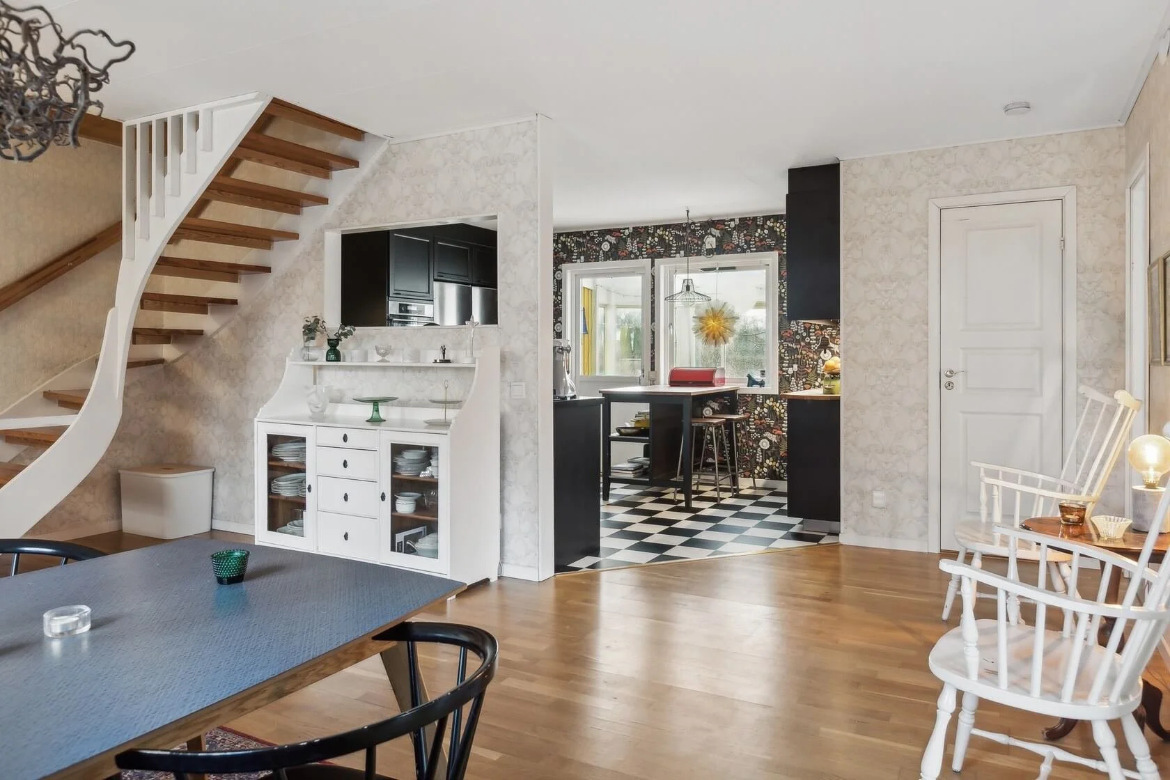 Living room with a staircase, dining table with chairs, and a kitchen area in the background.