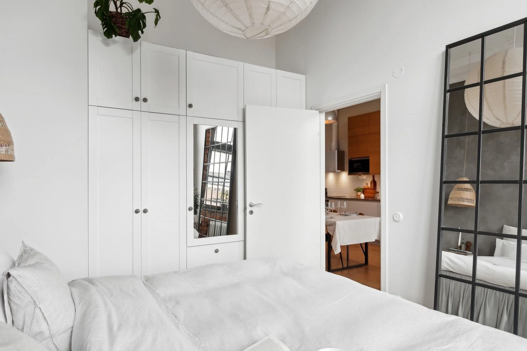 Bedroom with white bedding, built-in white wardrobe, mirror, and a large black-framed window reflecting the bedroom and part of the living space