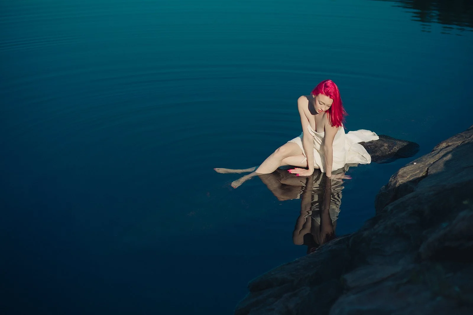 Woman with vibrant red hair sitting on rocks in water, wearing a white dress, looking down with her hands touching the water, and her reflection visible.