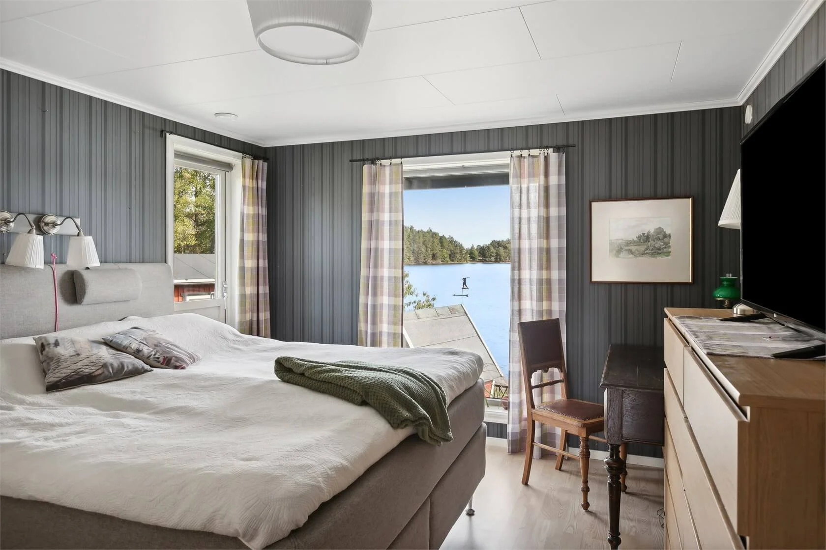 A bedroom with a large bed, a window showing a lake view, and a dresser with a TV on top.
