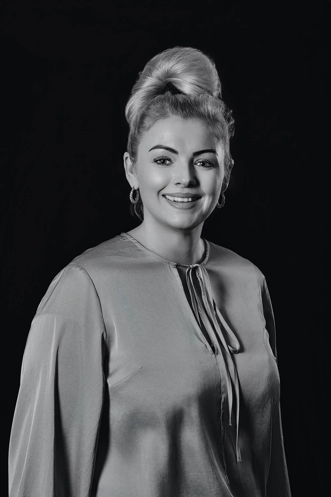 Black and white portrait of a smiling woman with styled hair, wearing earrings and a blouse with a tie at the neckline, against a dark background.