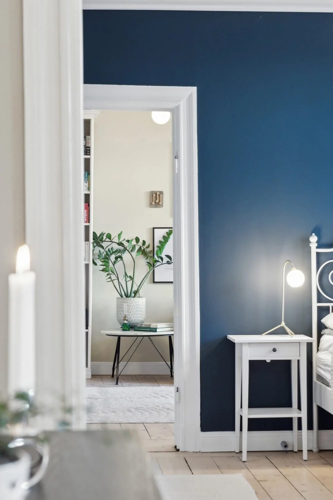 View of a bedroom with a white bed frame, a white bedside table with a reading lamp, a blue accent wall, and a doorway leading to a living space with a potted plant, a small round table, and wall art.