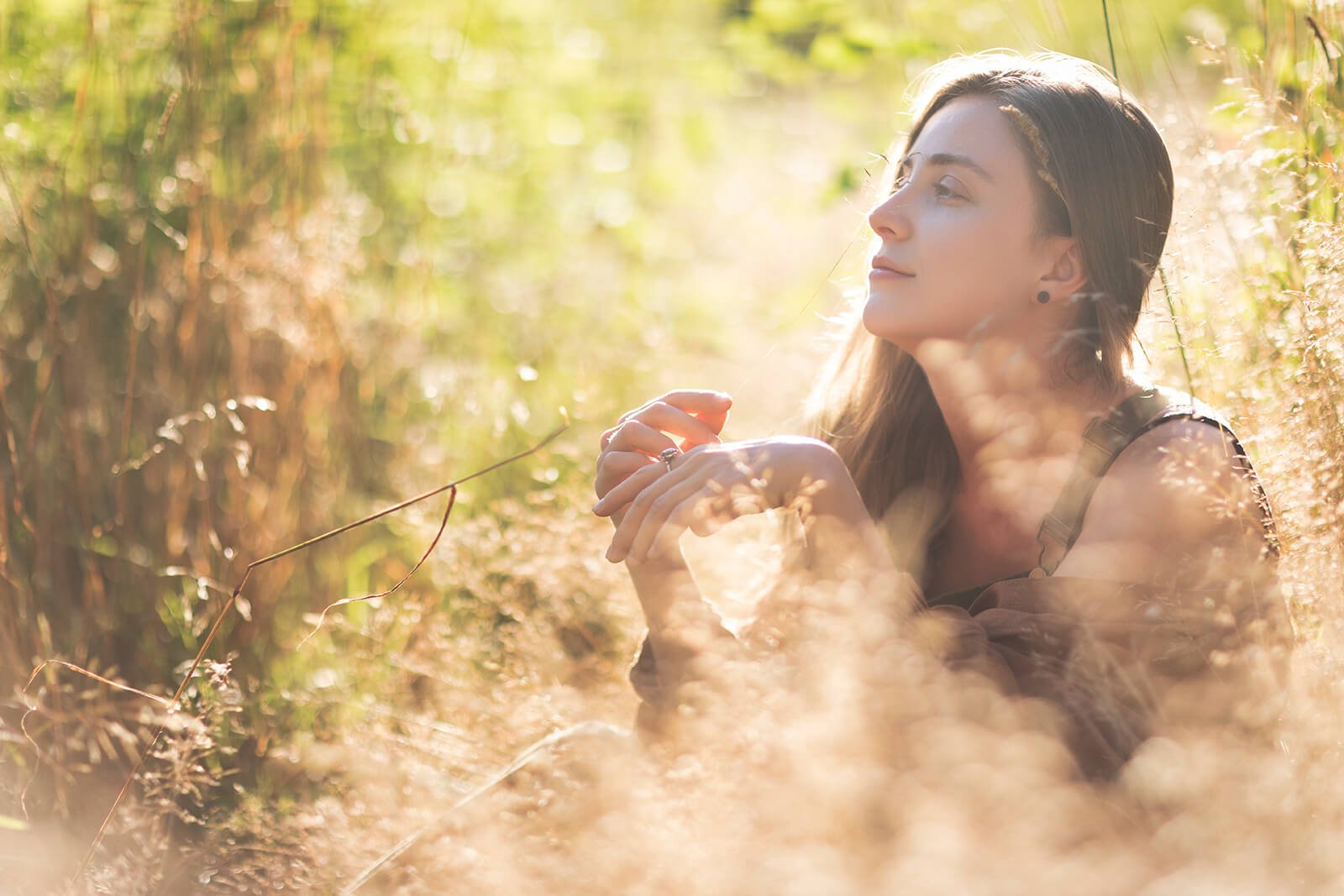 A woman sitting in the grass surrounded by tall dry plants and foliage, enjoying sunlight with a peaceful expression.
