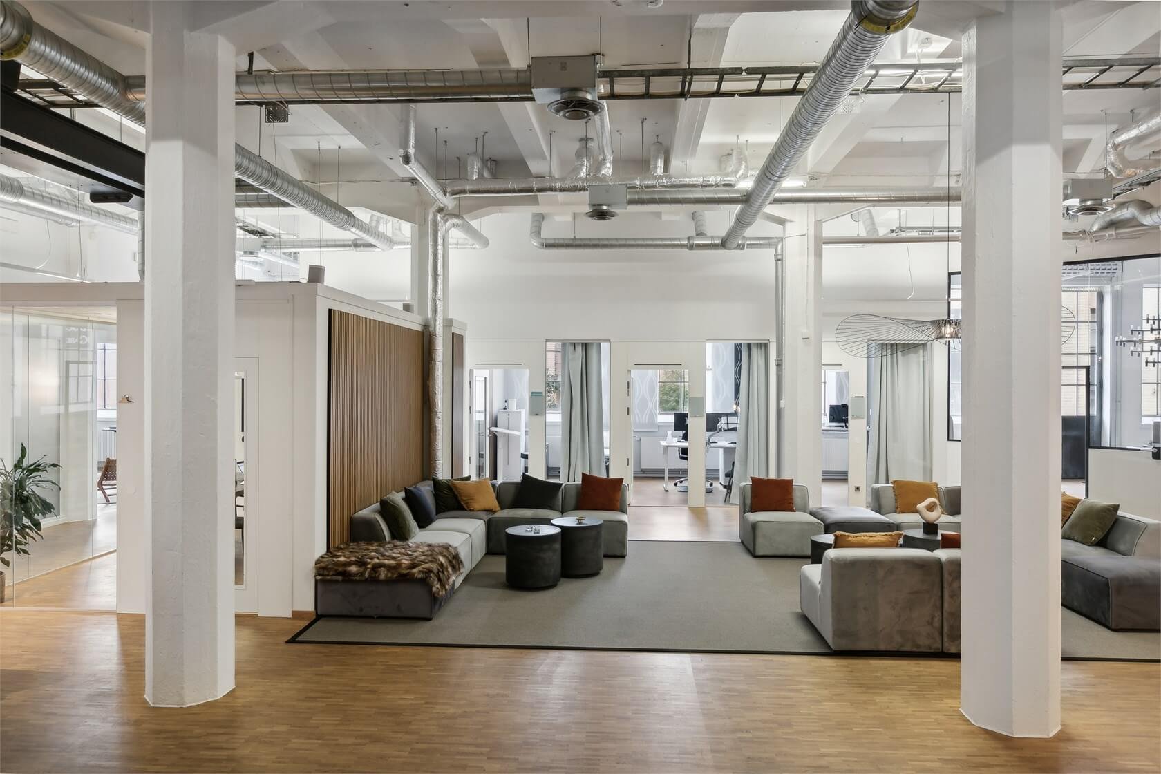 Modern office lobby with gray couches, pillows, black and brown accents, wooden and gray flooring, large windows, and visible office areas in the background.
