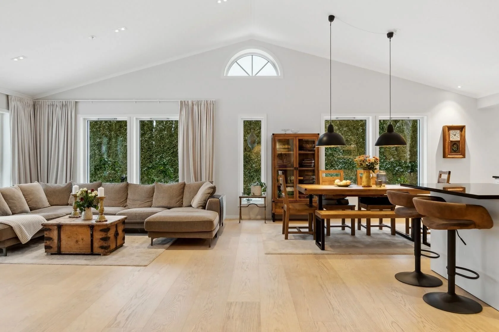 Open-concept living and dining area with large windows, beige sectional sofa, wooden coffee table, dining table with chairs, pendant lights, and kitchen bar stools.