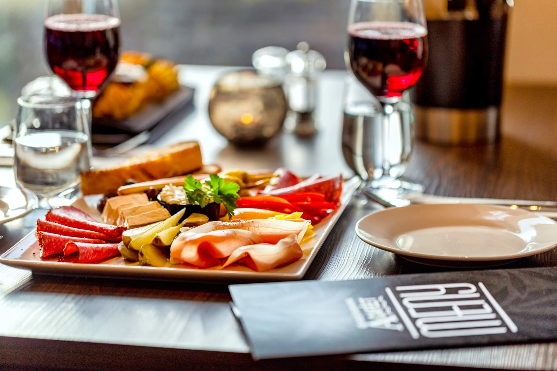 A charcuterie board with various slices of meats, cheese, pickles, and garnishes on a table set with wine glasses, water glasses, and plates at a dining table.