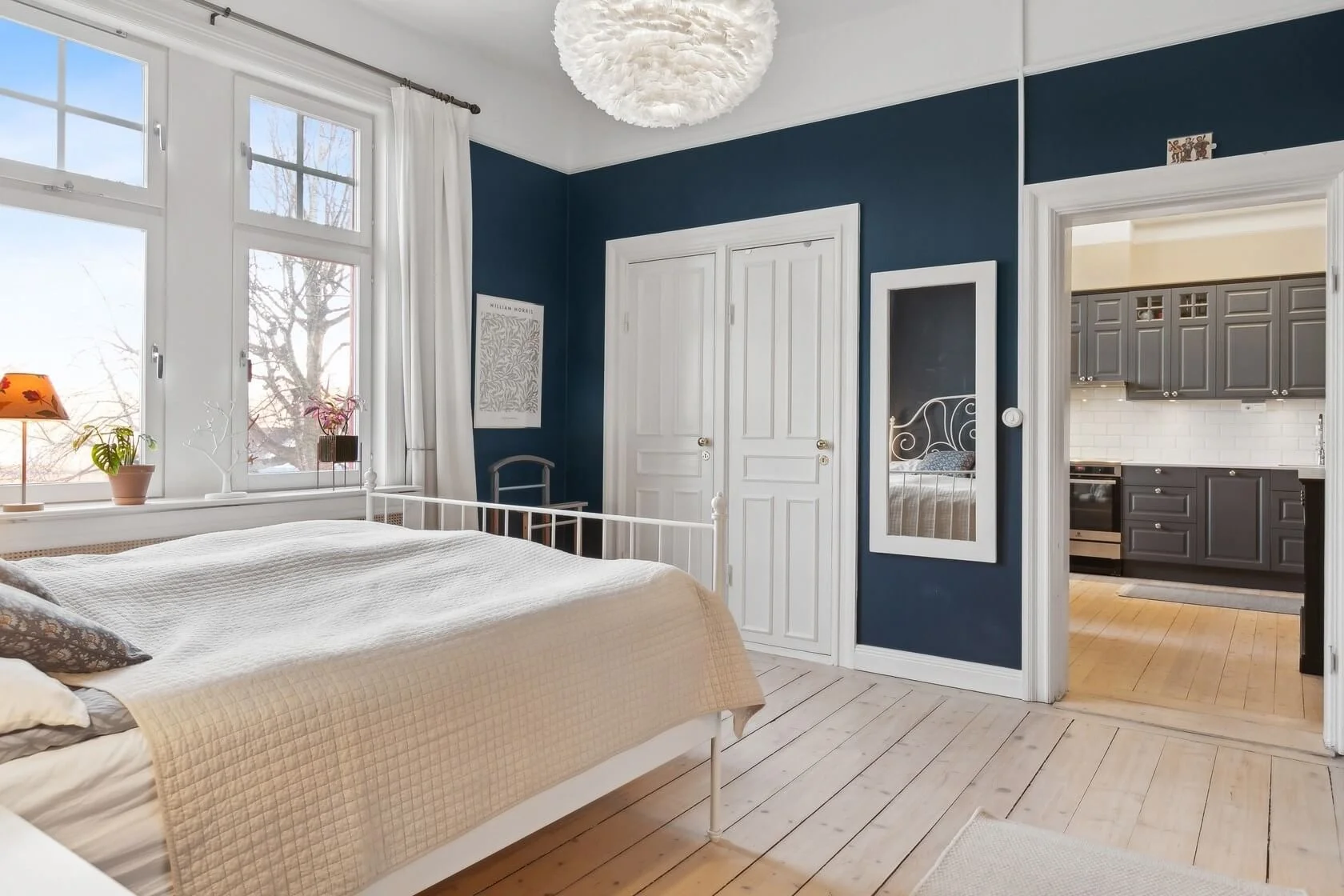 A bedroom with a white bed, beige quilt, and patterned pillows, large windows with white curtains, blue accent wall, full-length mirror, and view into a kitchen with gray cabinets.