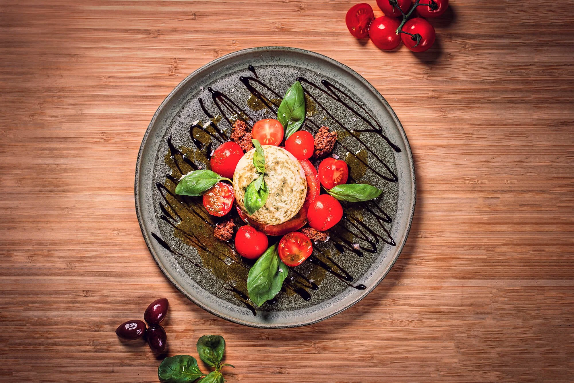 Caprese salad on a gray plate with cherry tomatoes, basil leaves, mozzarella, and a drizzle of balsamic glaze, placed on a wooden surface with additional tomatoes and olives nearby.