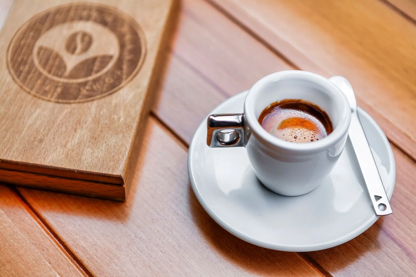 A cup of espresso on a white saucer with a silver spoon, placed on a wooden table near a wooden menu or coaster.