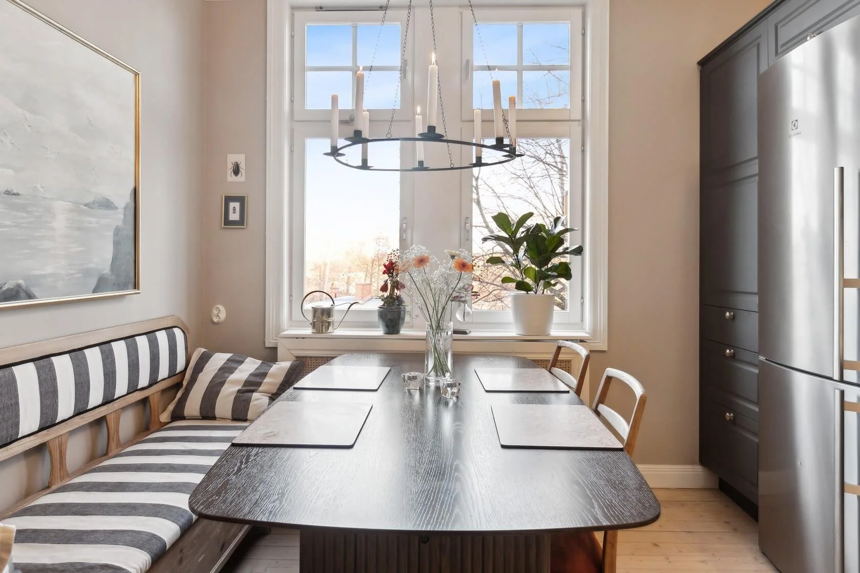 A dining area with a dark wooden table set with four place mats, a bench with black and white striped cushions on the left, three chairs on the right, and a vase of flowers in the center. Large windows with clear sky and trees outside are in the back