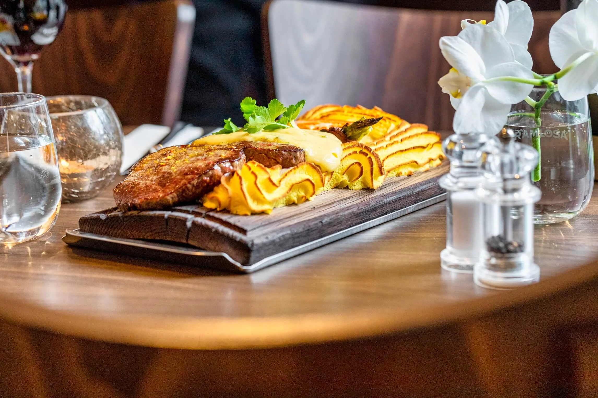 Steak with mashed potatoes, gravy, asparagus, and sliced yellow vegetables served on a wooden board, on a table with glasses of water, a flower in a glass vase, and salt and pepper shakers.