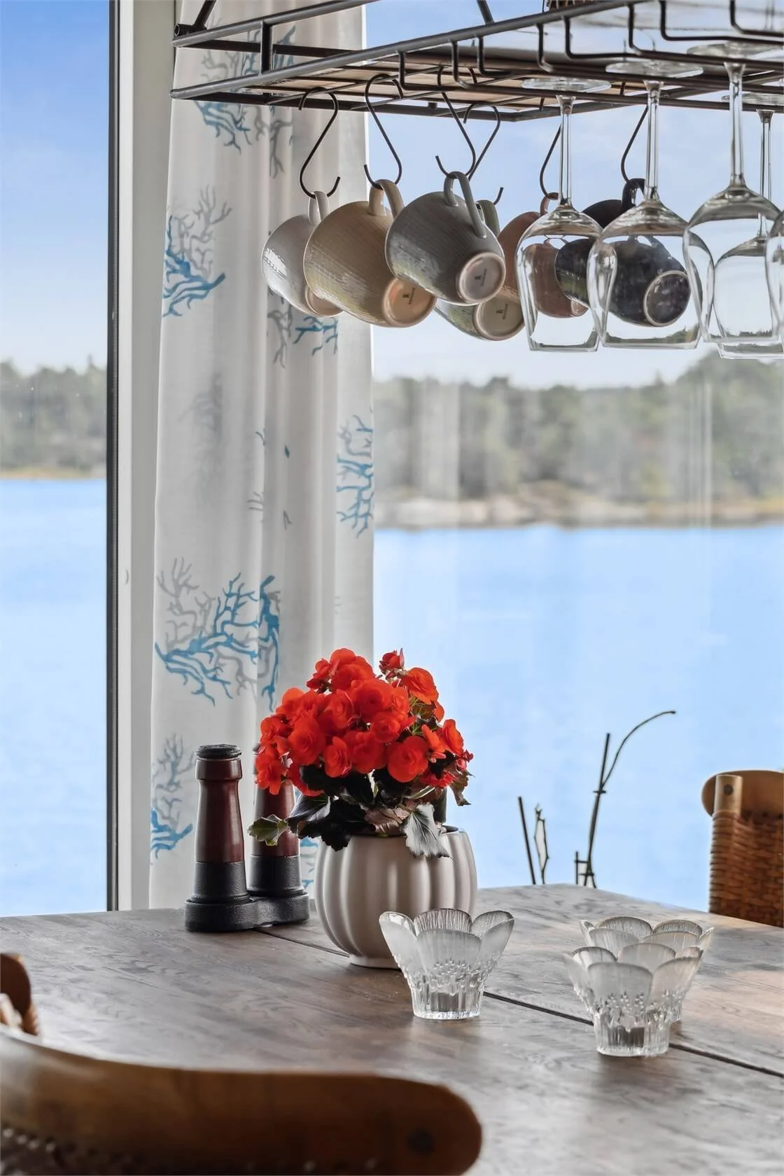 A wooden dining table near a window with a view of a lake and trees, decorated with a vase of red flowers, glassware, and salt and pepper shakers, with a hanging rack of cups and glasses above.