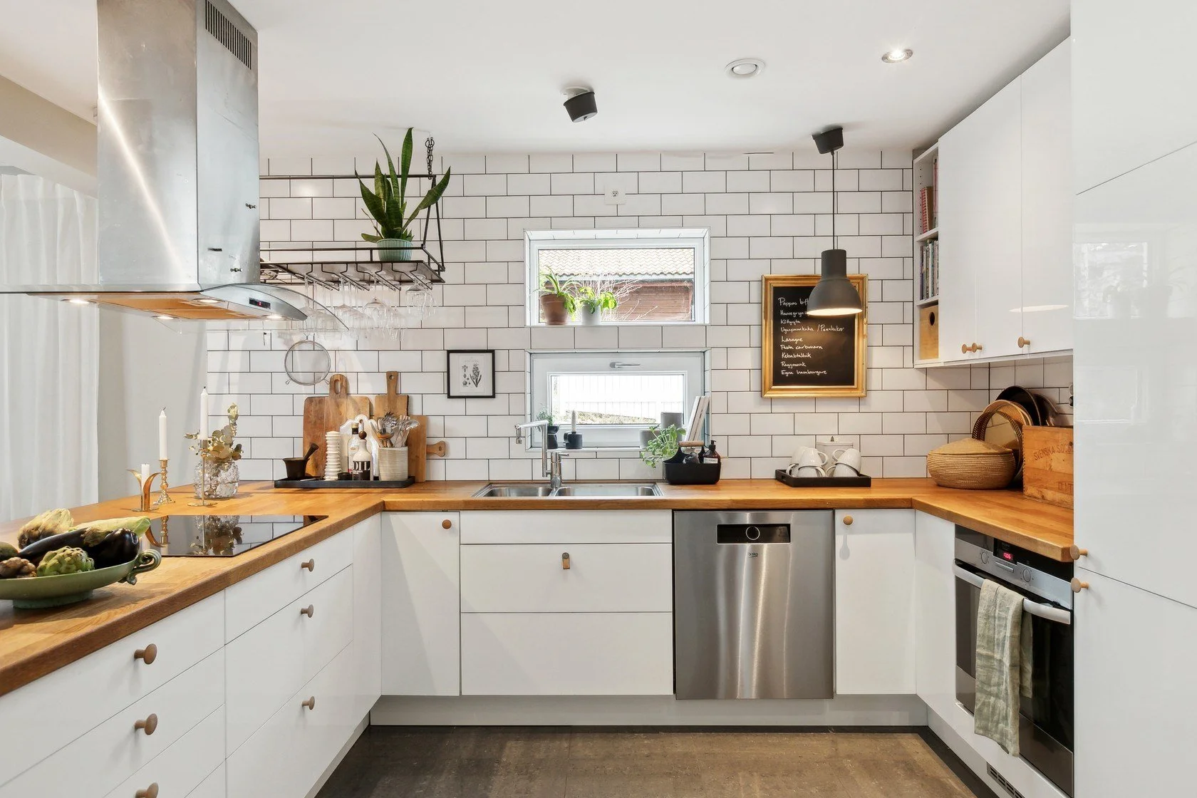 Modern kitchen with white cabinets, wooden countertops, a two-sink setup, and white subway tile backsplash. There are various kitchen items, plants on the windowsill, and a black chalkboard menu on the wall.