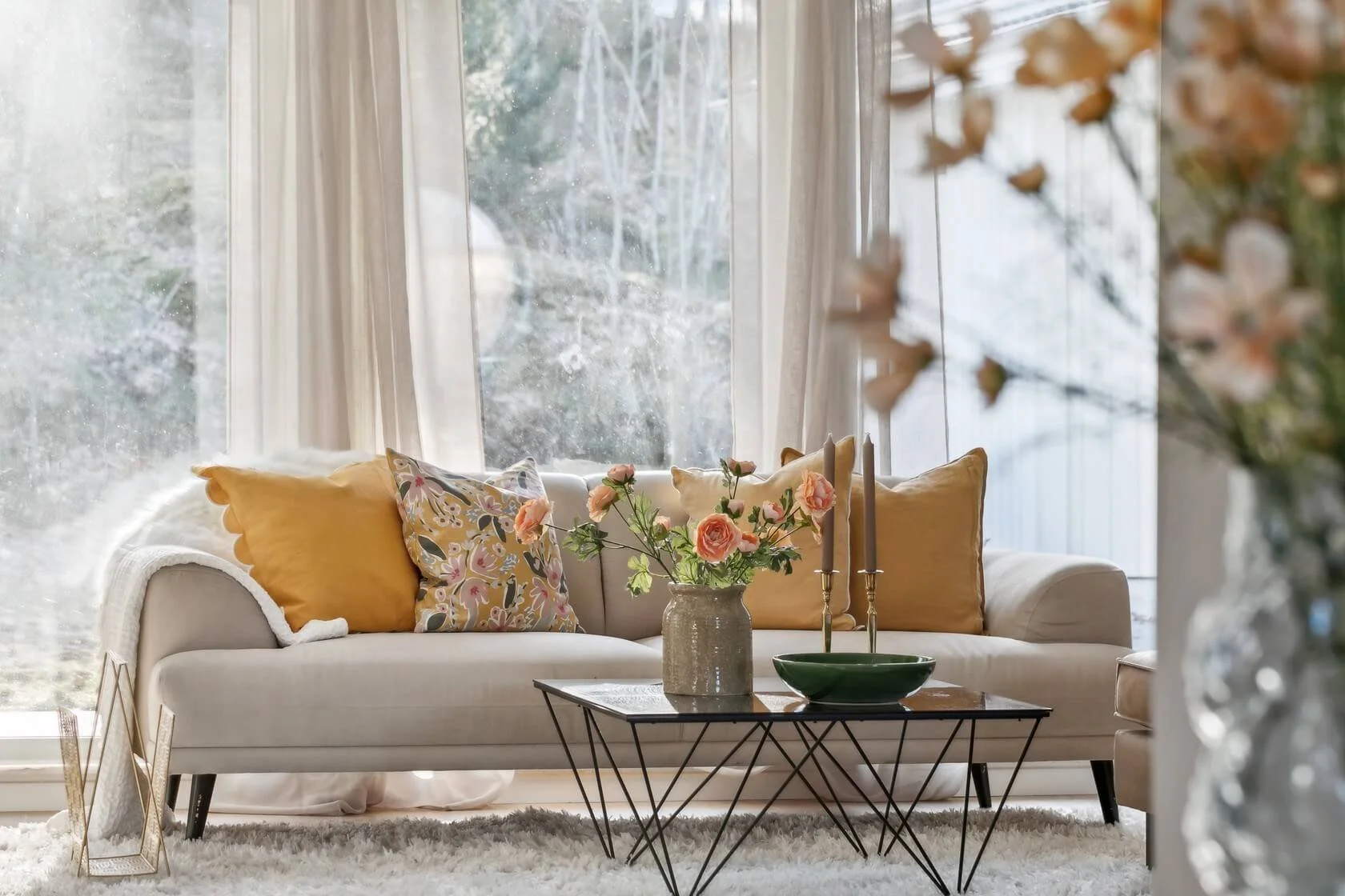 Living room with a white sofa set against large windows, decorated with yellow and patterned cushions, with a coffee table holding pink flowers in a beige vase, and two candlesticks, in front of a white fluffy rug.