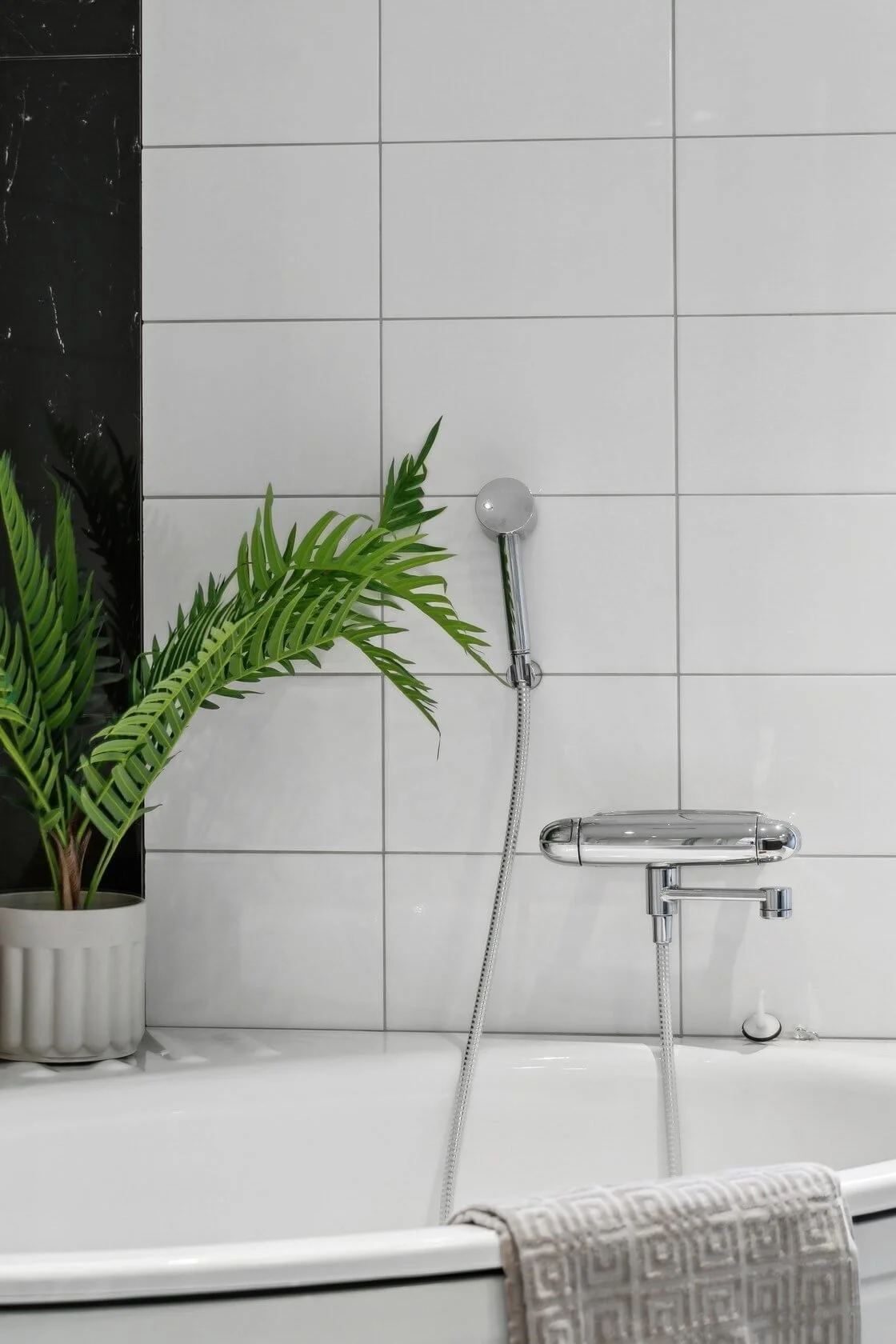 A bathroom with white tiled walls, a potted plant on the edge of a bathtub, and a chrome showerhead and faucet.
