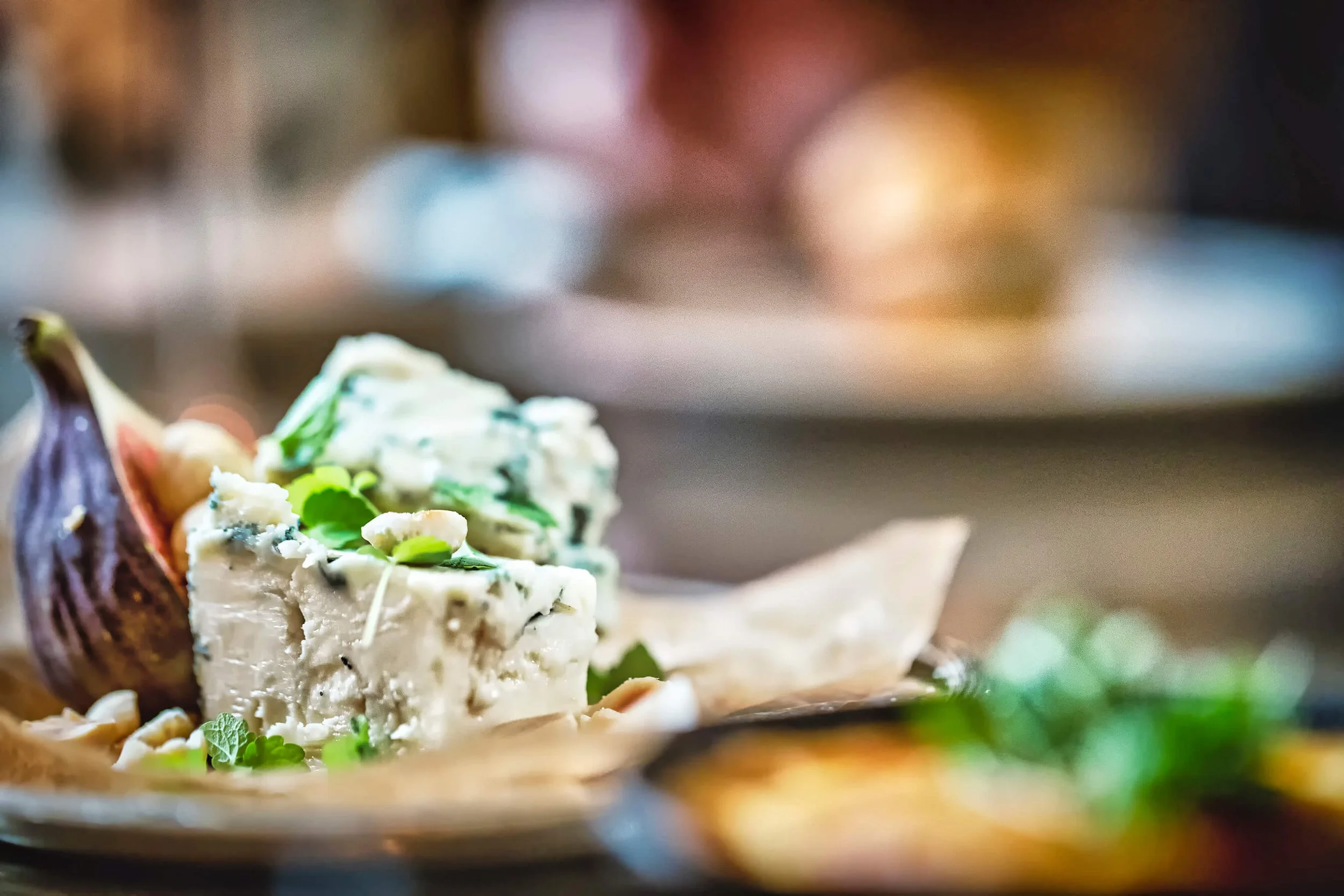 Close-up of a wedge of blue cheese garnished with green herbs, with a roasted garlic bulb and other cheeses blurred in the background.