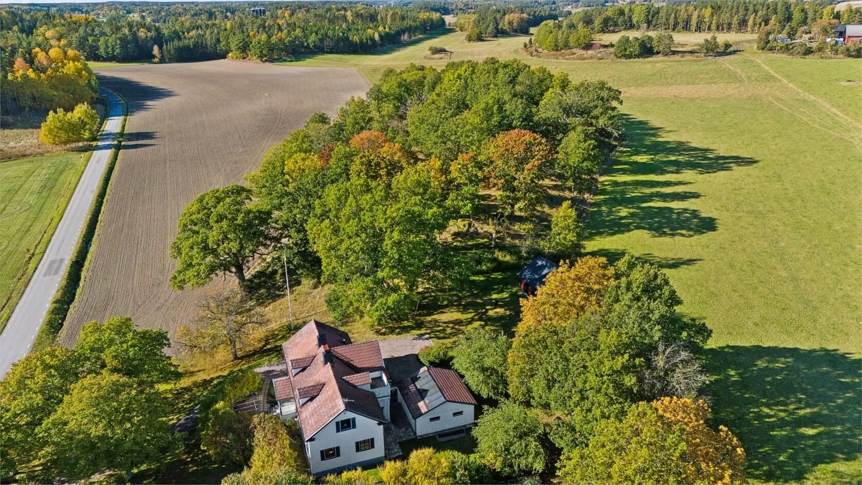 Aerial view of a rural landscape featuring a white house with a brown roof, surrounded by trees with green and fall-colored leaves, adjacent to open fields and farmland, with a road running along the edge of the fields.