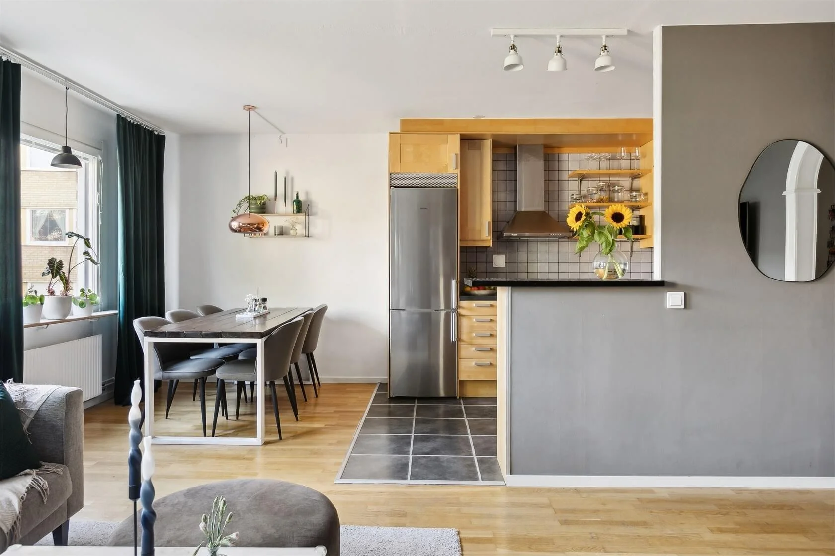 Open living room and kitchen area with a dining table, gray chairs, a gray accent wall with a round mirror, and a kitchen with wooden cabinets, a stainless steel refrigerator, and sunflower centerpiece.