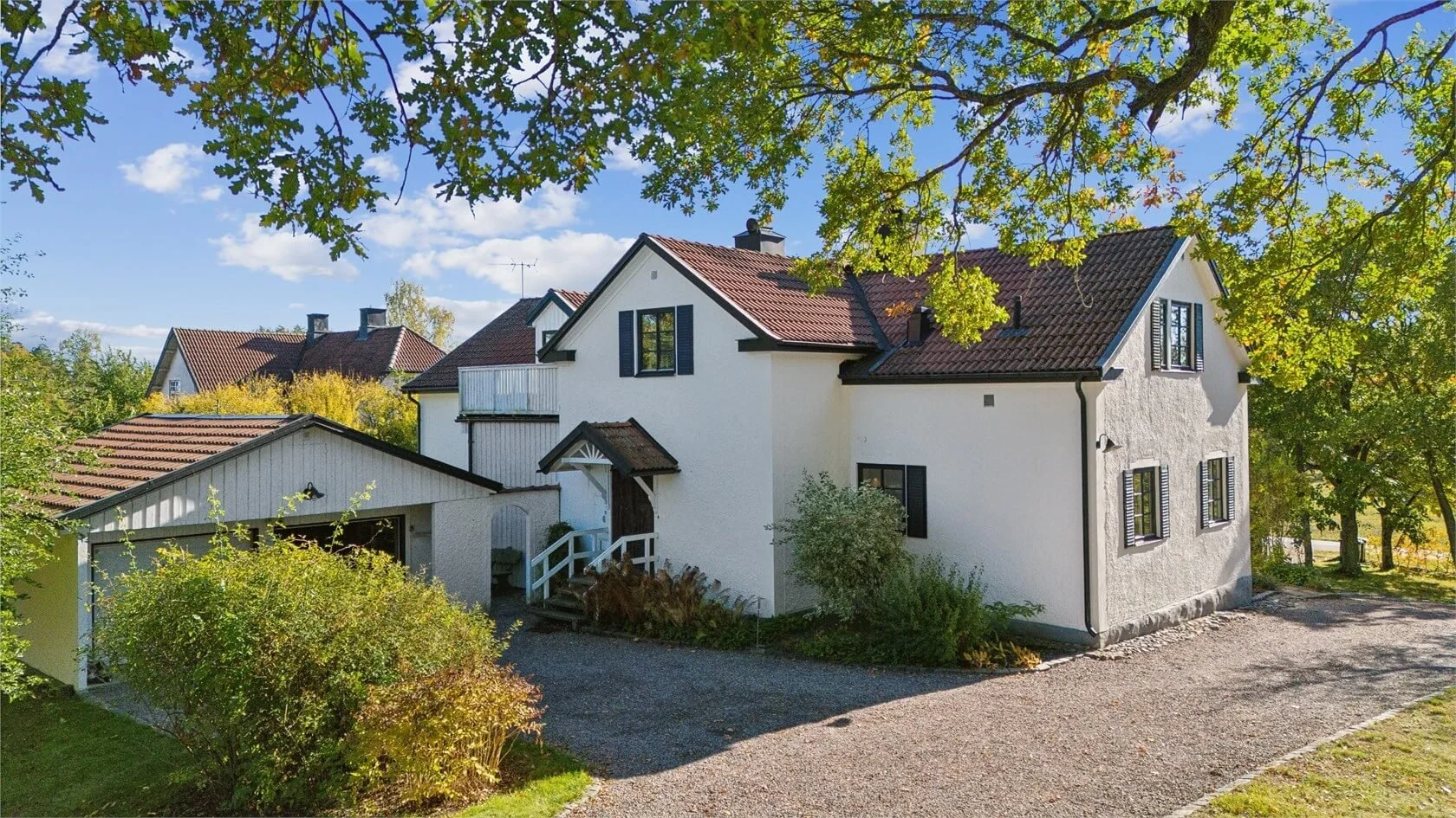 A white house with black window shutters and a red tiled roof, surrounded by green trees and shrubs, with a gravel driveway in front, under a blue sky.