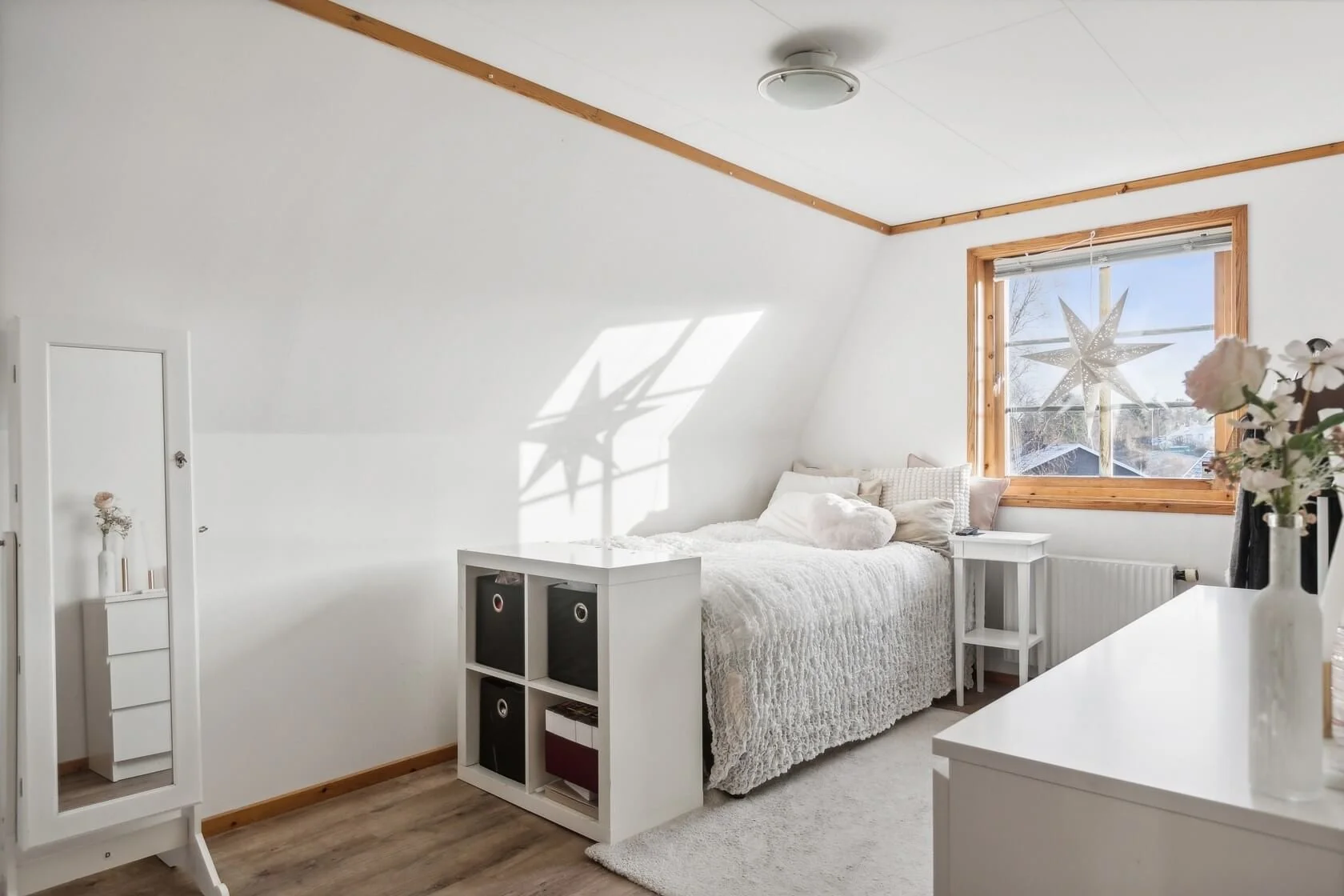 A cozy bedroom with a white bed, window with wood trim, and star-shaped decoration in the window. White furniture and minimal decor create a bright, airy atmosphere.