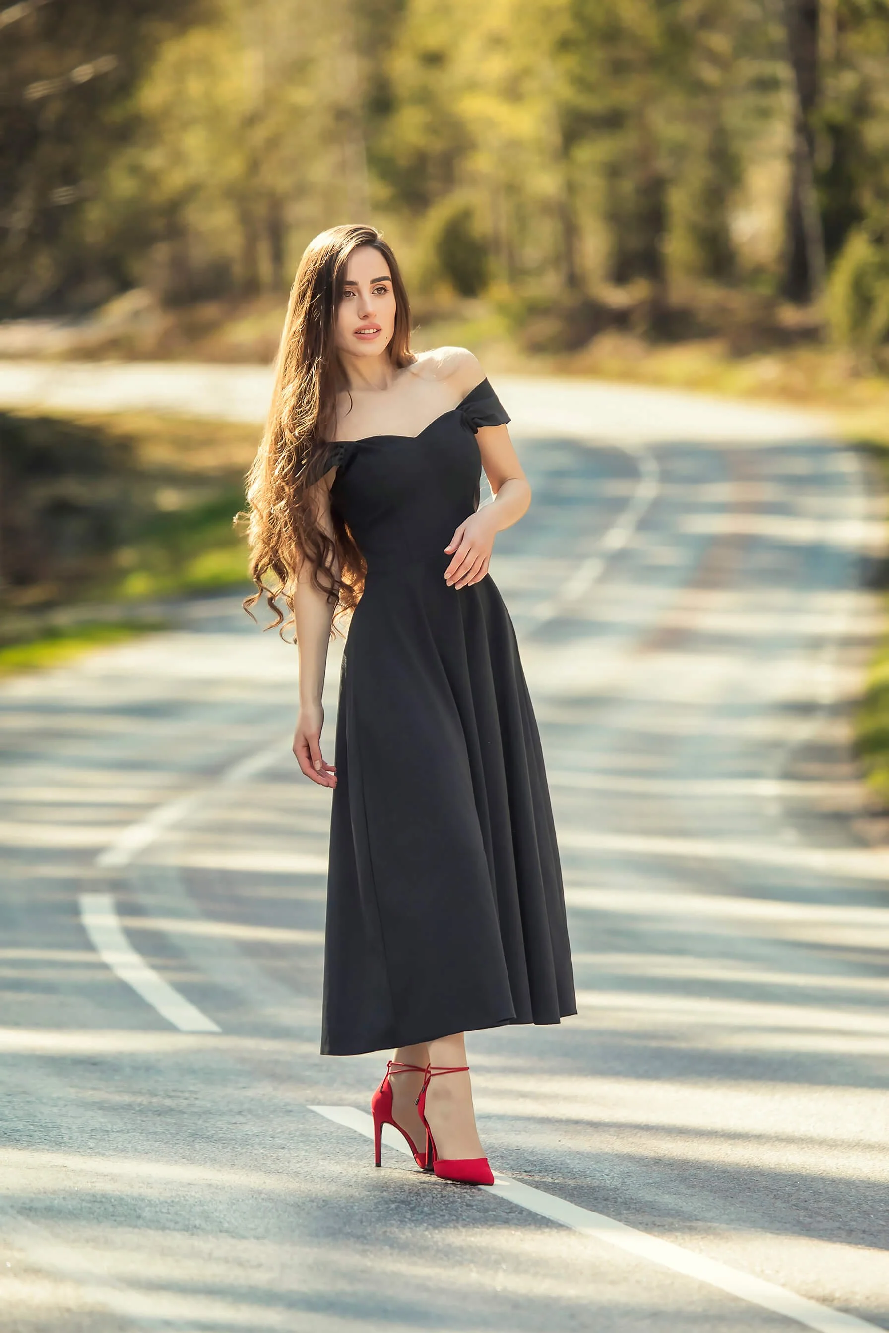 A woman in a black off-shoulder dress and red high heels standing on a winding road surrounded by trees.