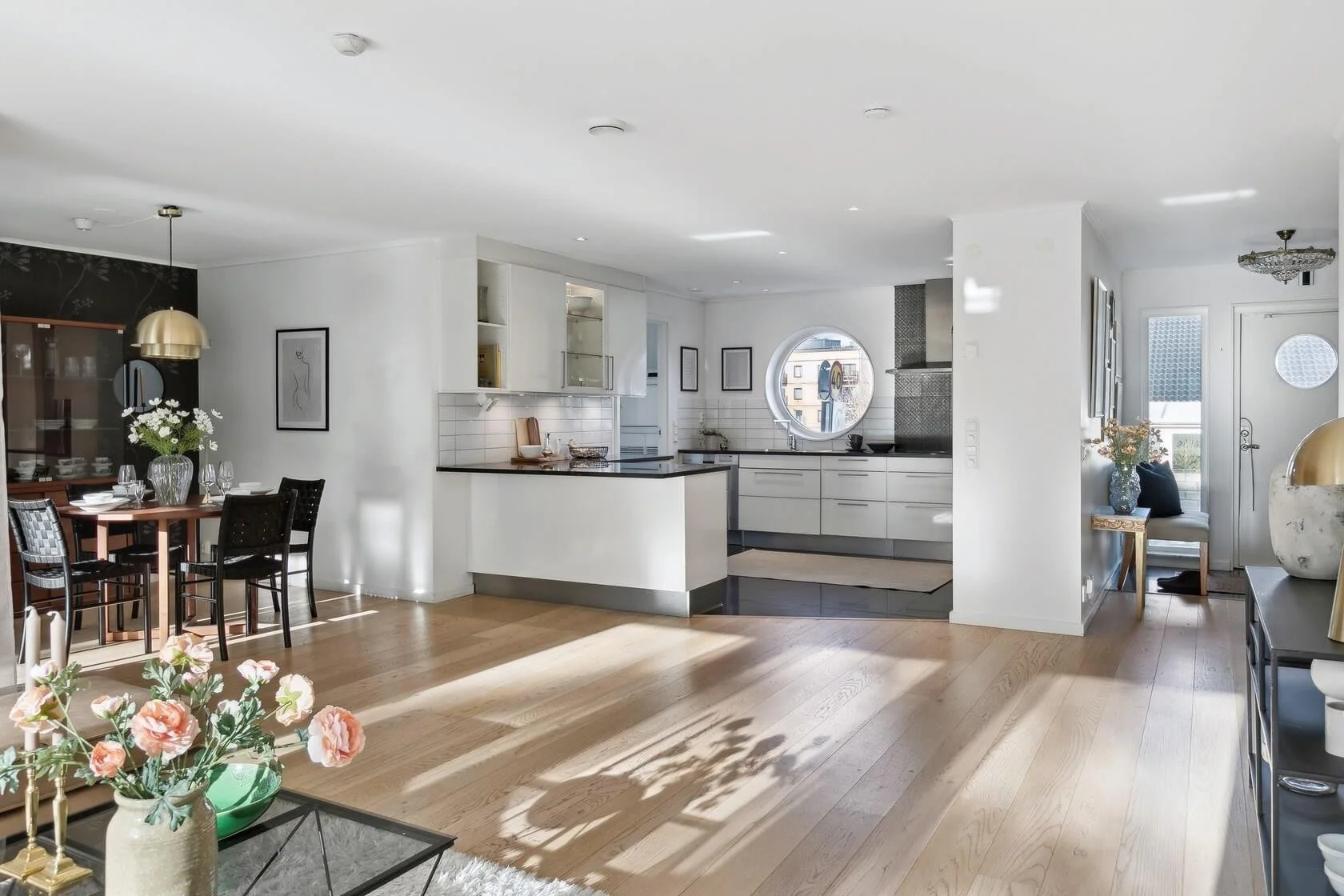Open-concept modern living space with a dining area and kitchen featuring white cabinets, black countertops, and a circular window, with hardwood flooring and decor including flowers and framed artwork.