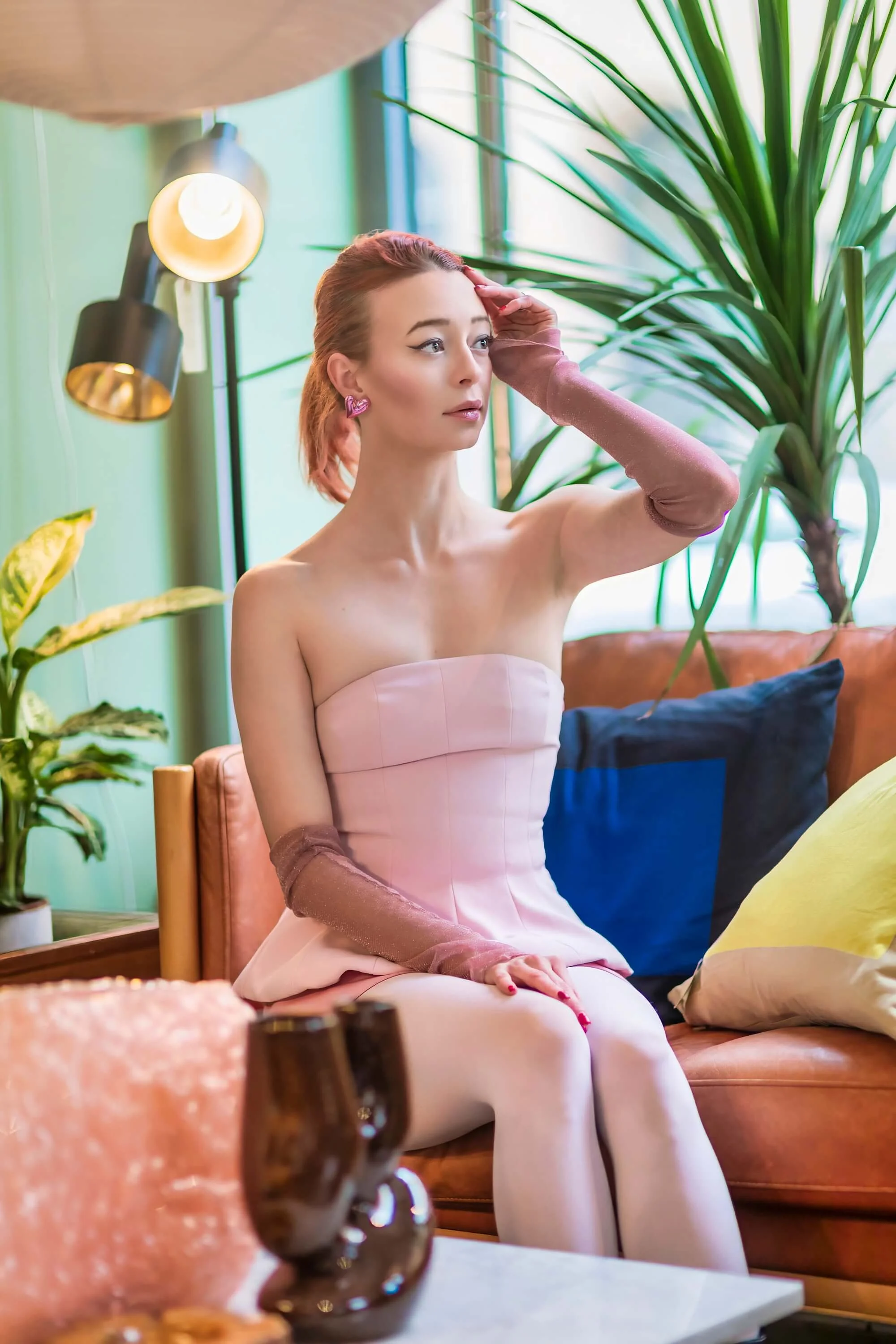 A woman with pink hair, wearing a strapless pink dress and pink gloves, sitting on a sofa with colorful pillows, in a room with green walls, large plants, and standing lamps.