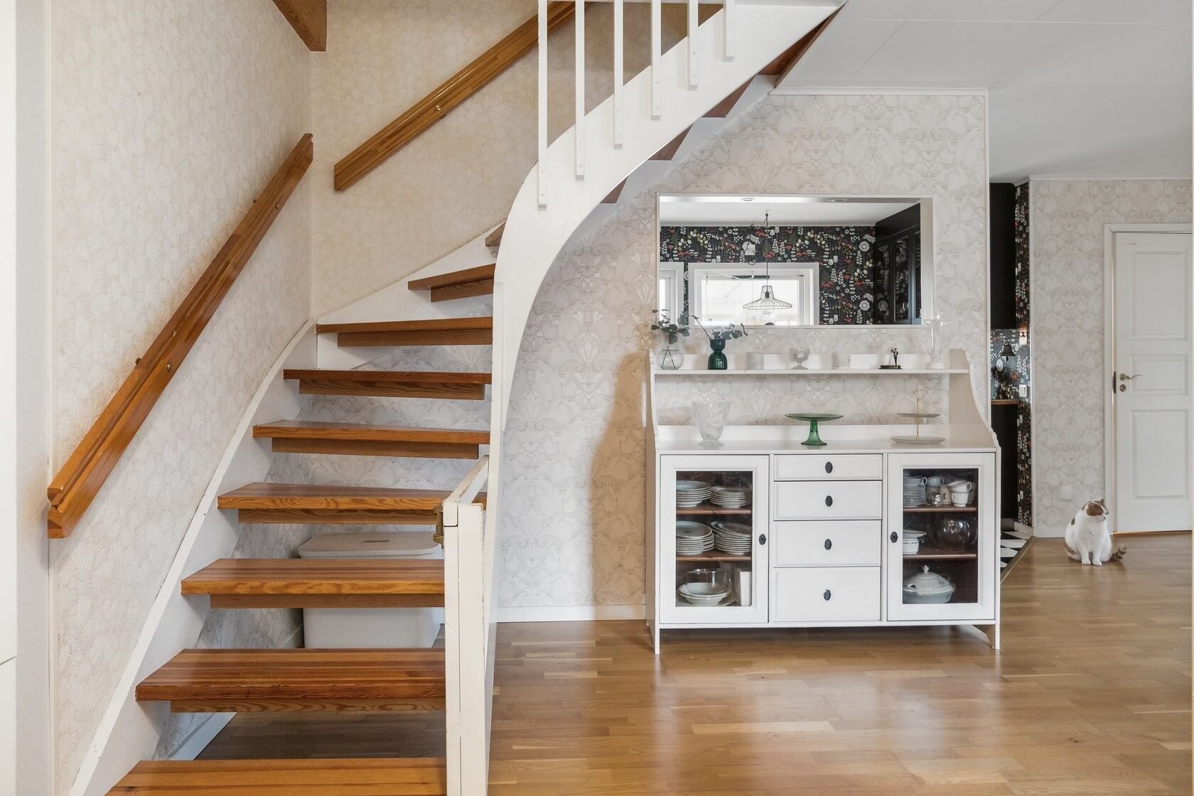 Interior of a home showing a curved wooden staircase on the left, a white cabinet with glass doors filled with dishes in the center, a decorative mirror above it, and a cat sitting on the wooden floor near a white door on the right. The walls have pa