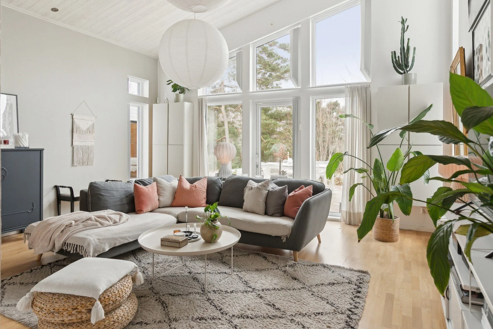 Bright living room with large windows, white walls, gray sofa with pink and beige pillows, potted plants, round coffee table with decorative items, woven ottoman, modern decor, and light wood flooring.