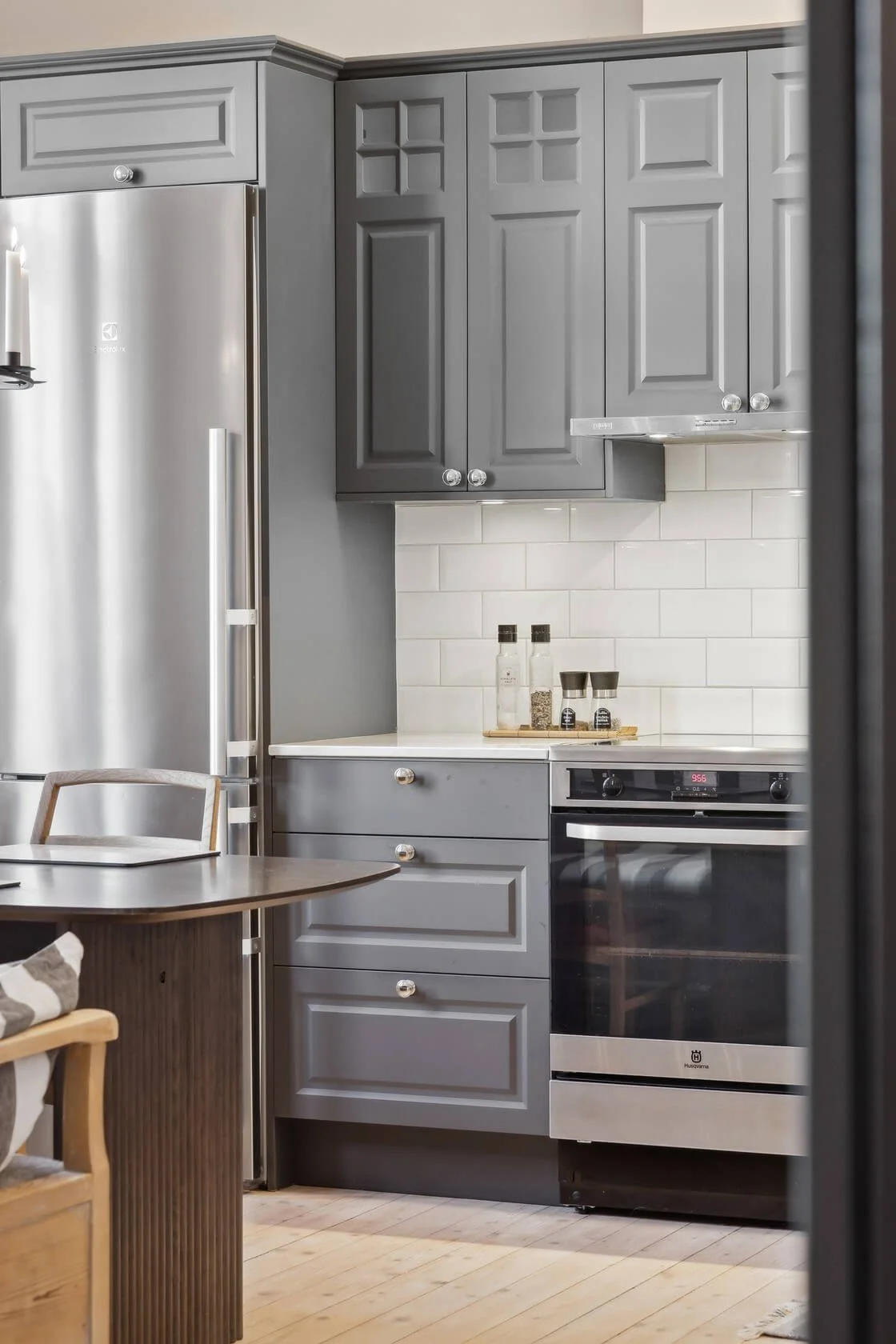 A modern kitchen with gray cabinets, a stainless steel refrigerator, a white countertop, white subway tile backsplash, and a black and stainless steel oven. There are spice jars and salt and pepper shakers on the counter, with a small wooden table an