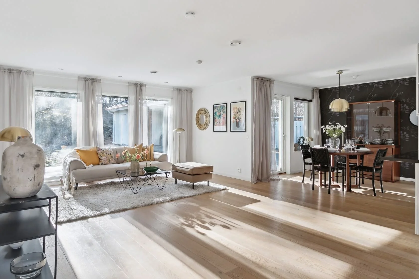 Bright living room with large windows, white sofa decorated with yellow and patterned pillows, glass coffee table, beige ottoman, black metal side table with a large ceramic vase, artwork on white wall, black accent wall with floral pattern, wooden d