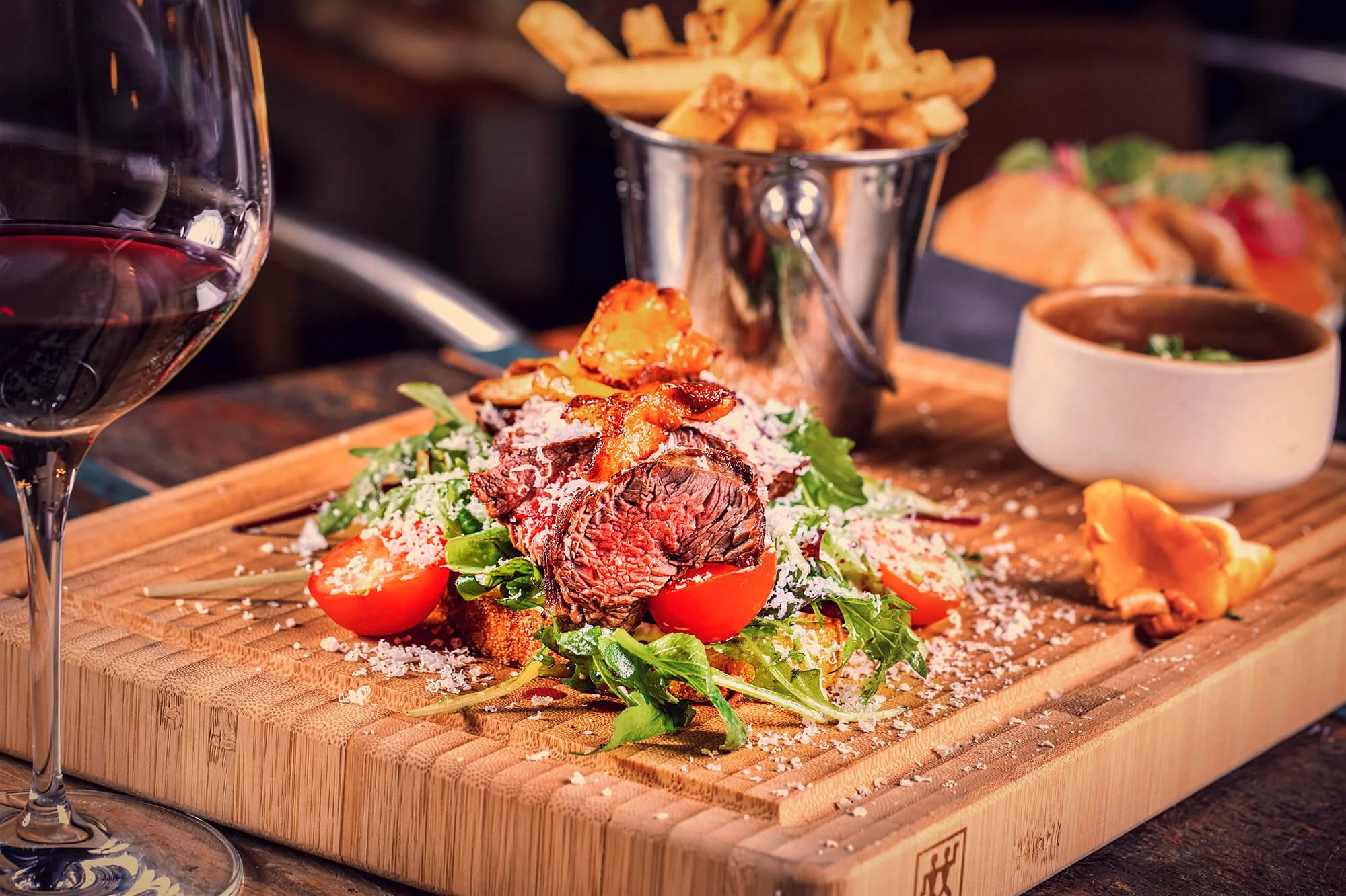 A meal on a wooden serving board featuring a sliced steak salad with cherry tomatoes, crispy bacon, and greens, with a side of French fries in a metal bucket, a bowl of soup, and a glass of red wine.