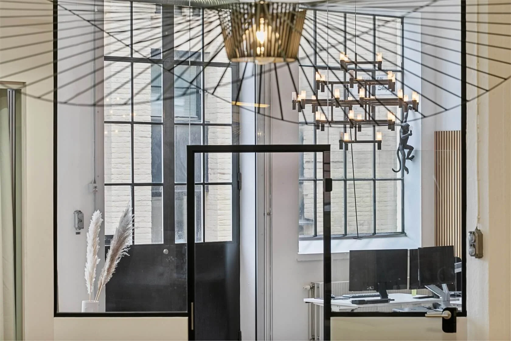 Modern office space seen through a glass partition with black frames, featuring a white desk with dual monitors, a black office chair, decorative pampas grass in a white pot, a black overlapping square sculpture, and geometric chandeliers with candle