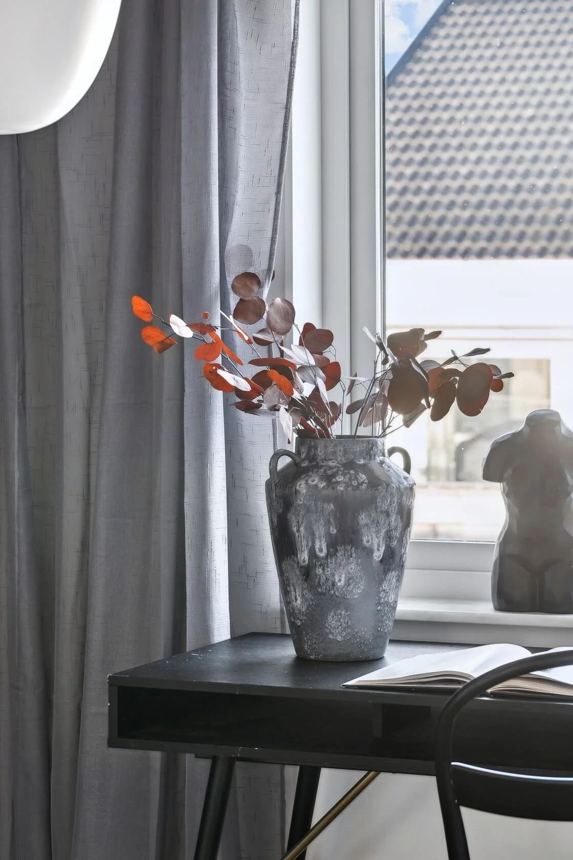 A gray ceramic vase with dusty pink and orange dried leaves, placed on a black desk near a window with gray curtains. A dog figurine and an open book are also on the desk.