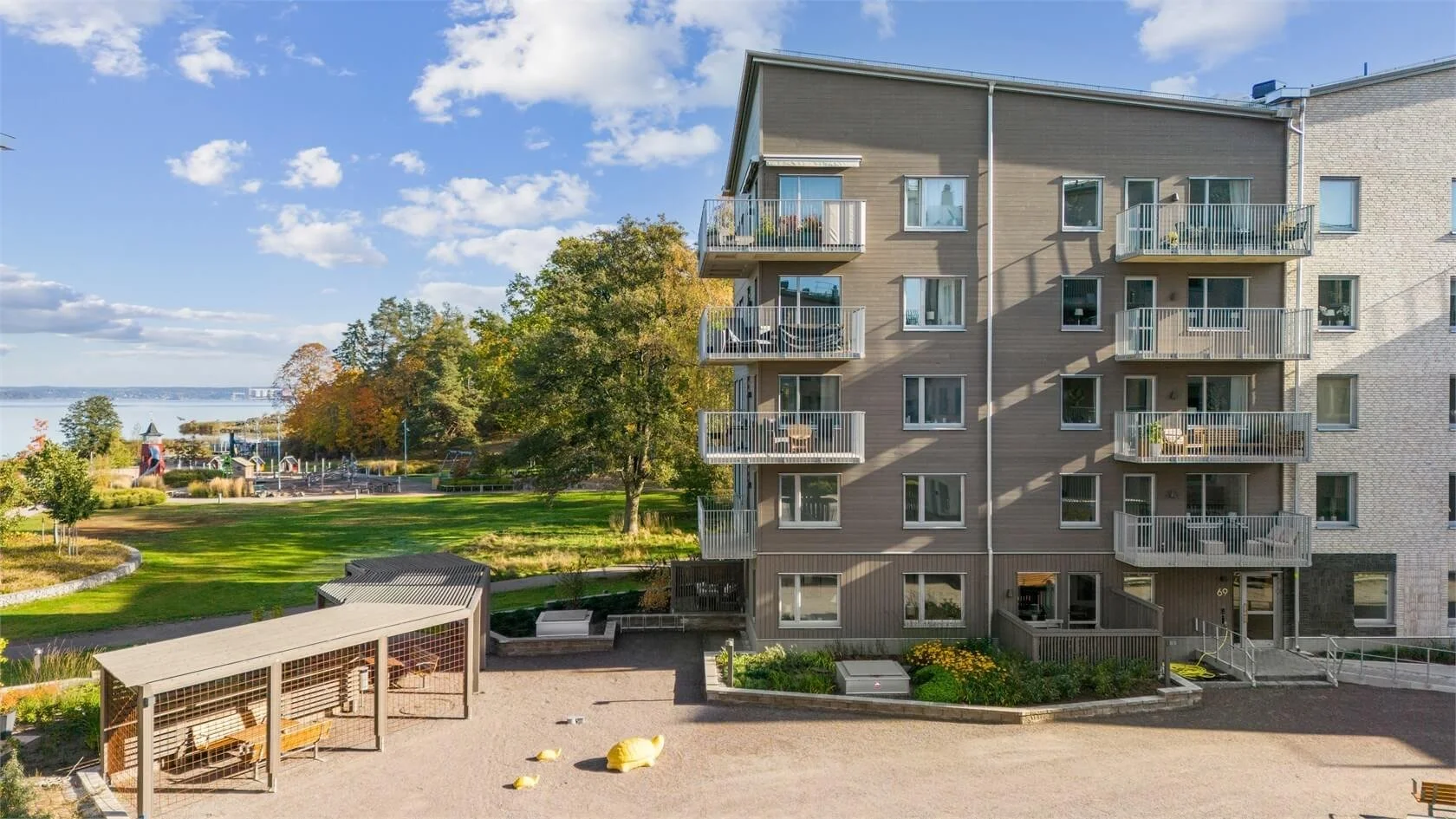 A multi-story apartment building with balconies, a park with trees and green grass, a playground with equipment, and a waterfront view with water and trees in the background.