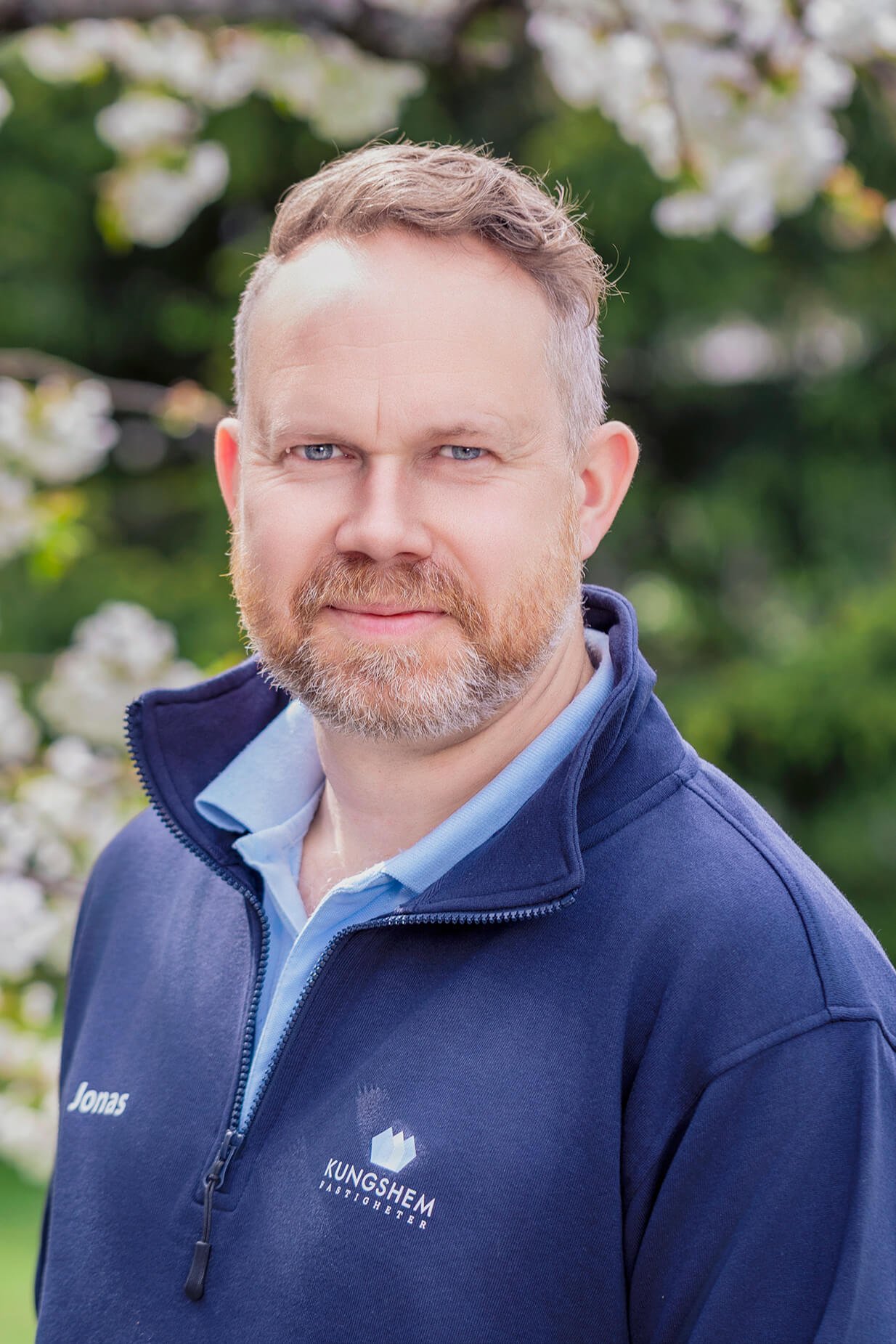 A man with short light brown hair, blue eyes, and a beard, wearing a navy blue quarter-zip pullover with a logo and text 'KUNG SHIEM PASTOR' and 'Jonas' on it, standing outdoors in front of green foliage and white flowers.