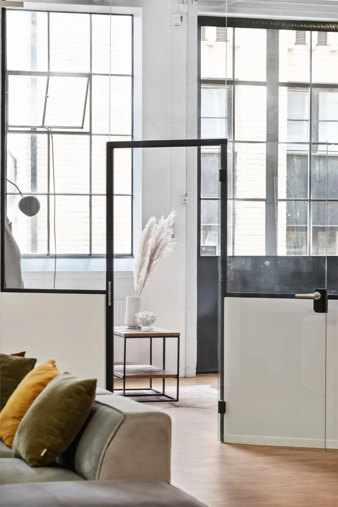 Modern living room with large industrial-style windows, a beige sofa with green and mustard cushions, a small wooden side table with a white vase of pampas grass, and black-framed glass doors.