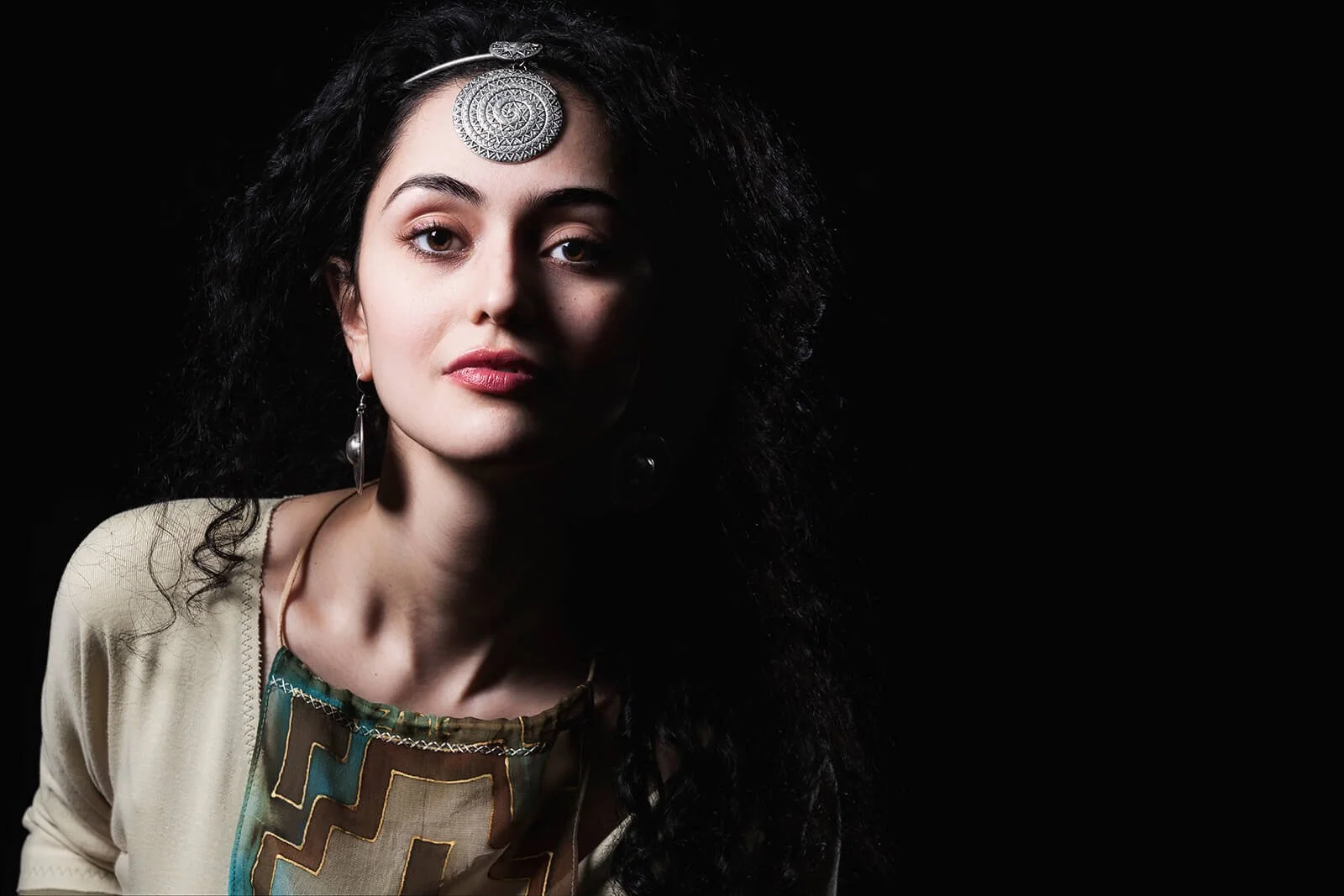 A woman with long, curly black hair styled in an voluminous manner, wearing a beige top with a colorful geometric pattern at the neckline, silver earrings, and a large silver pendant on her forehead, looking directly at the camera with a serious expr