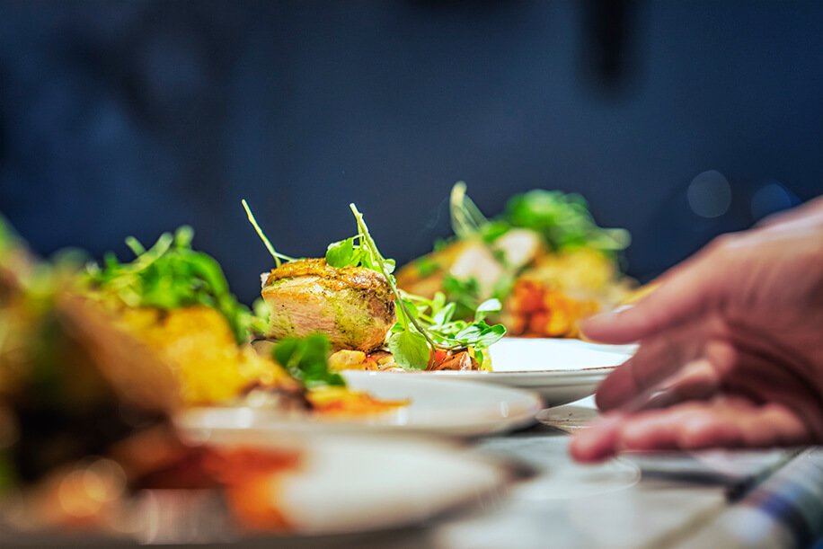 Close-up of a chef's hand plating a gourmet dish with greens and crusted meat on a white plate.