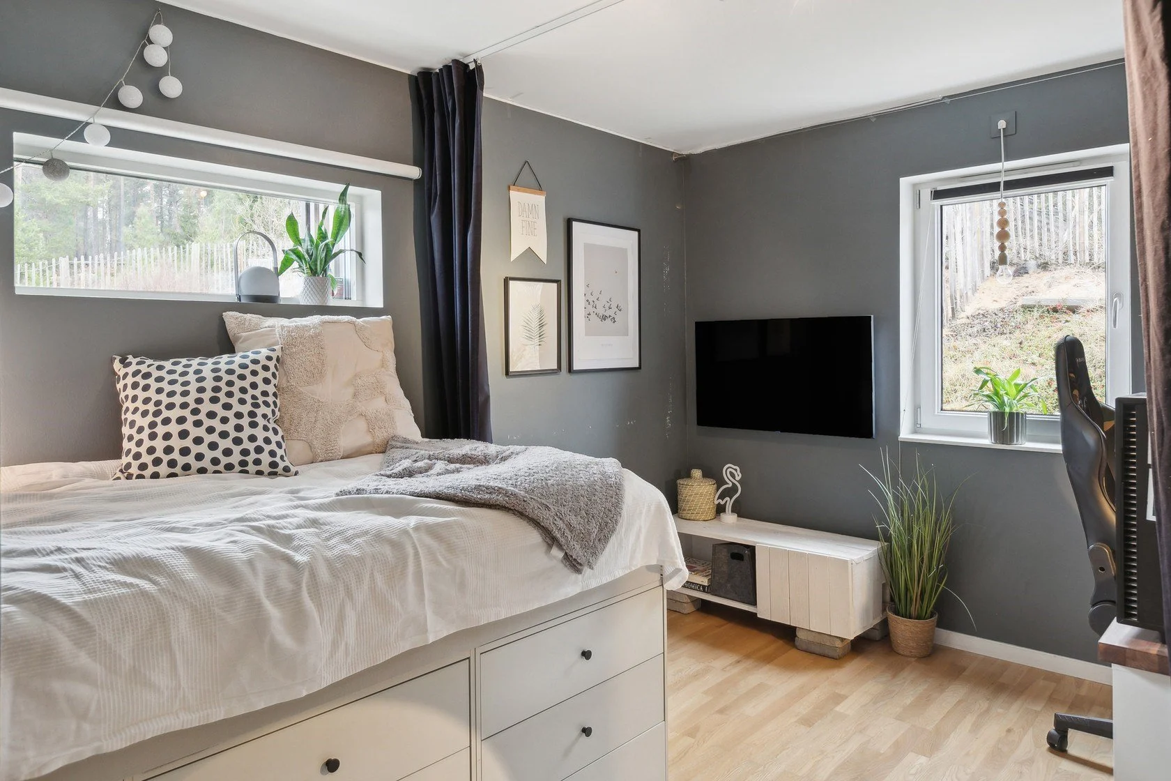 A bedroom with gray walls, a window above the bed with a plant and round white decorative items, a bed with white and patterned pillows, and a throw blanket. Art prints on the wall, a flat-screen TV, and a cabinet with decorative items and a plant ne