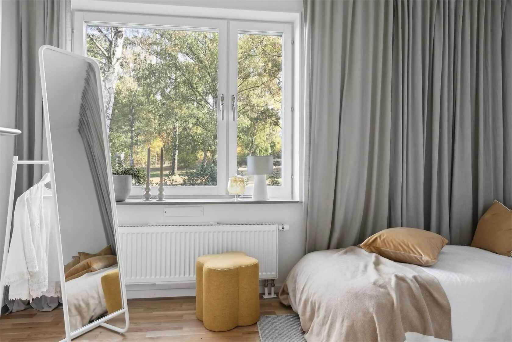 A bedroom with a large window showing trees outside, beige curtains, a white bed with beige pillows and blanket, a yellow ottoman, a full-length mirror, and a small table lamp.