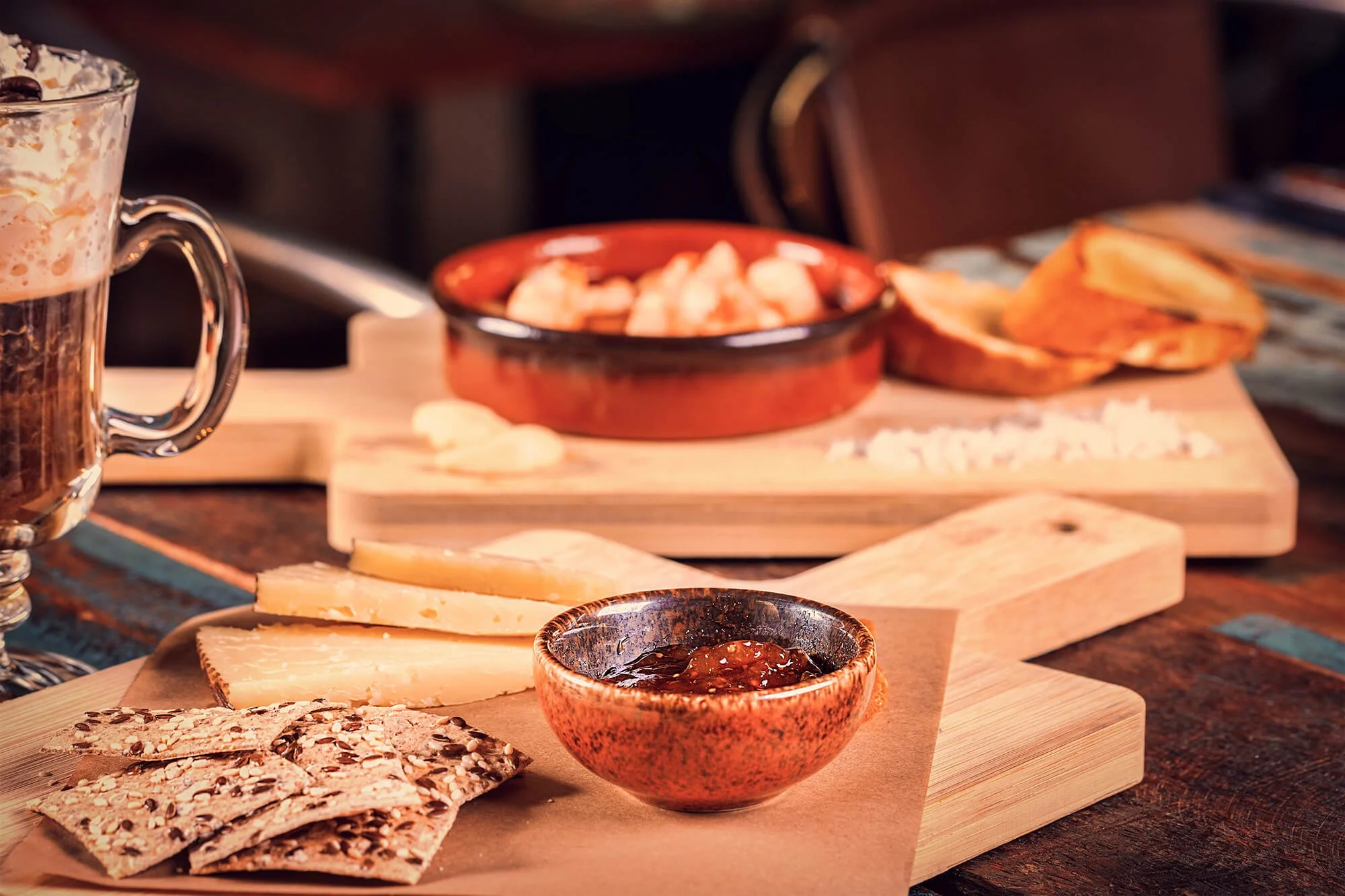 A cheese and snack platter on a wooden board with slices of cheese, seed crackers, and a bowl of jam, next to a glass of beer, on a rustic wooden table.