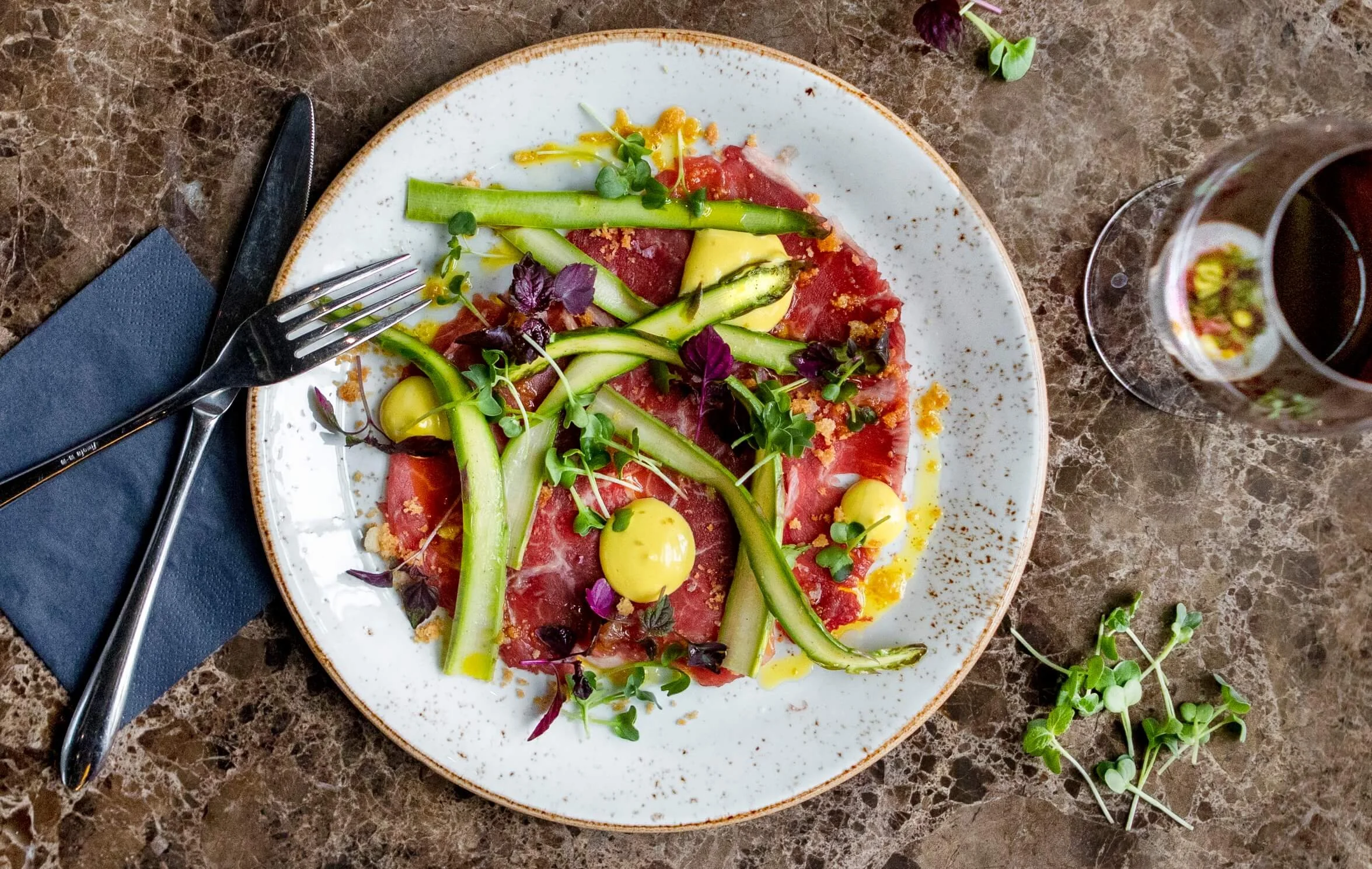 A salad with sliced roast beef, cucumber, microgreens, yellow sauce, and edible flowers on a white plate, with a glass of red wine nearby on a marble table.