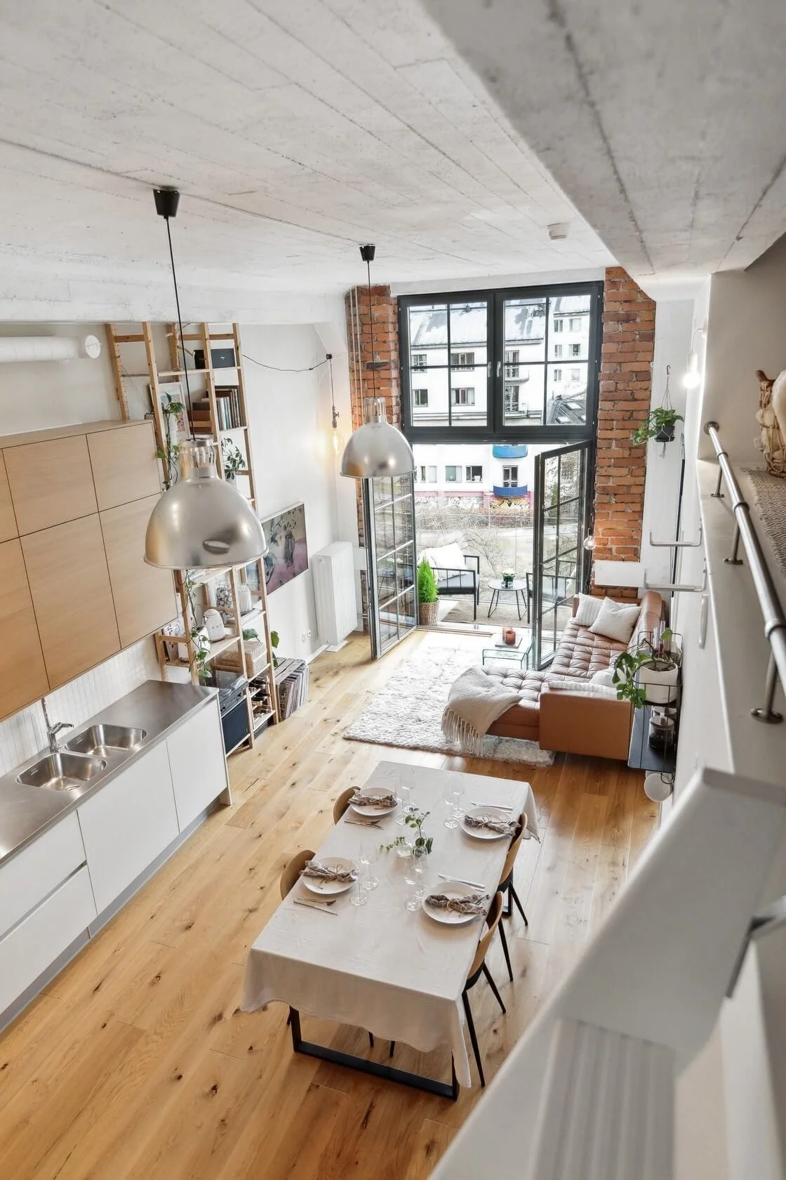 Interior view of a modern loft apartment with kitchen, dining area, and living room. Large windows open to a small balcony, with exposed brick and white walls, wood flooring, and contemporary decor.