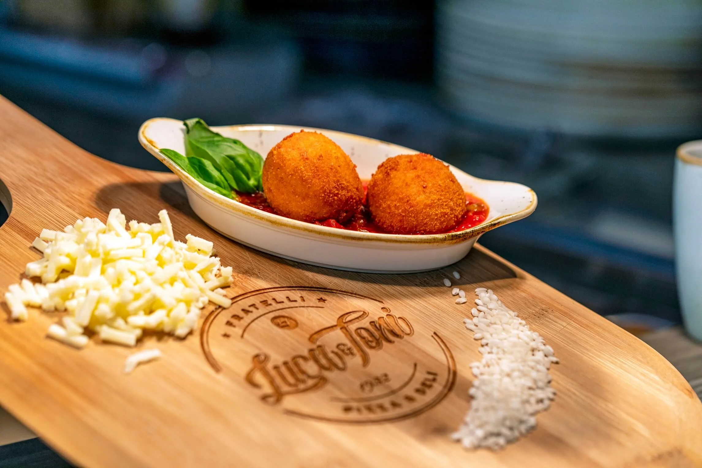 Two breaded fried balls on a white dish with sauce and basil leaves, accompanied by shredded cheese and rice on a wooden cutting board.