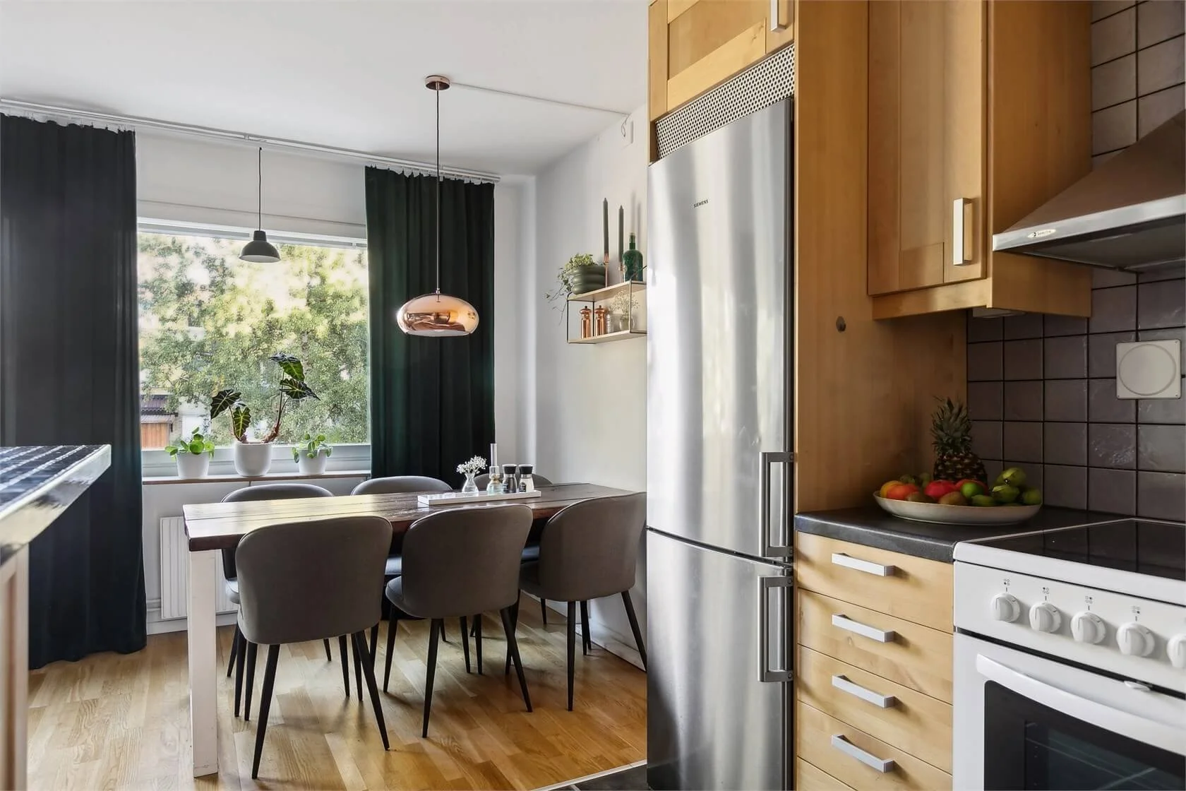 Kitchen with wooden cabinets, stainless steel refrigerator, black countertop, and a white stove. Dining area with a wooden table, six gray chairs, and a large window with three potted plants and dark curtains.